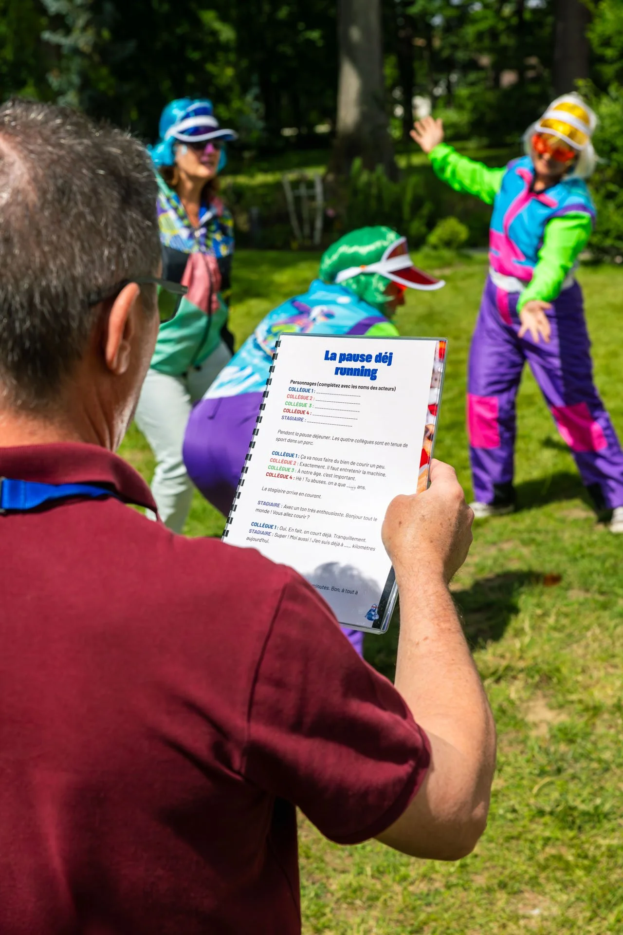 Un homme lit un script pour un jeu de rôle ou une activité en plein air, avec un groupe de personnes habillées comme des personnages de dessin animé, dans un parc ensoleillé.
