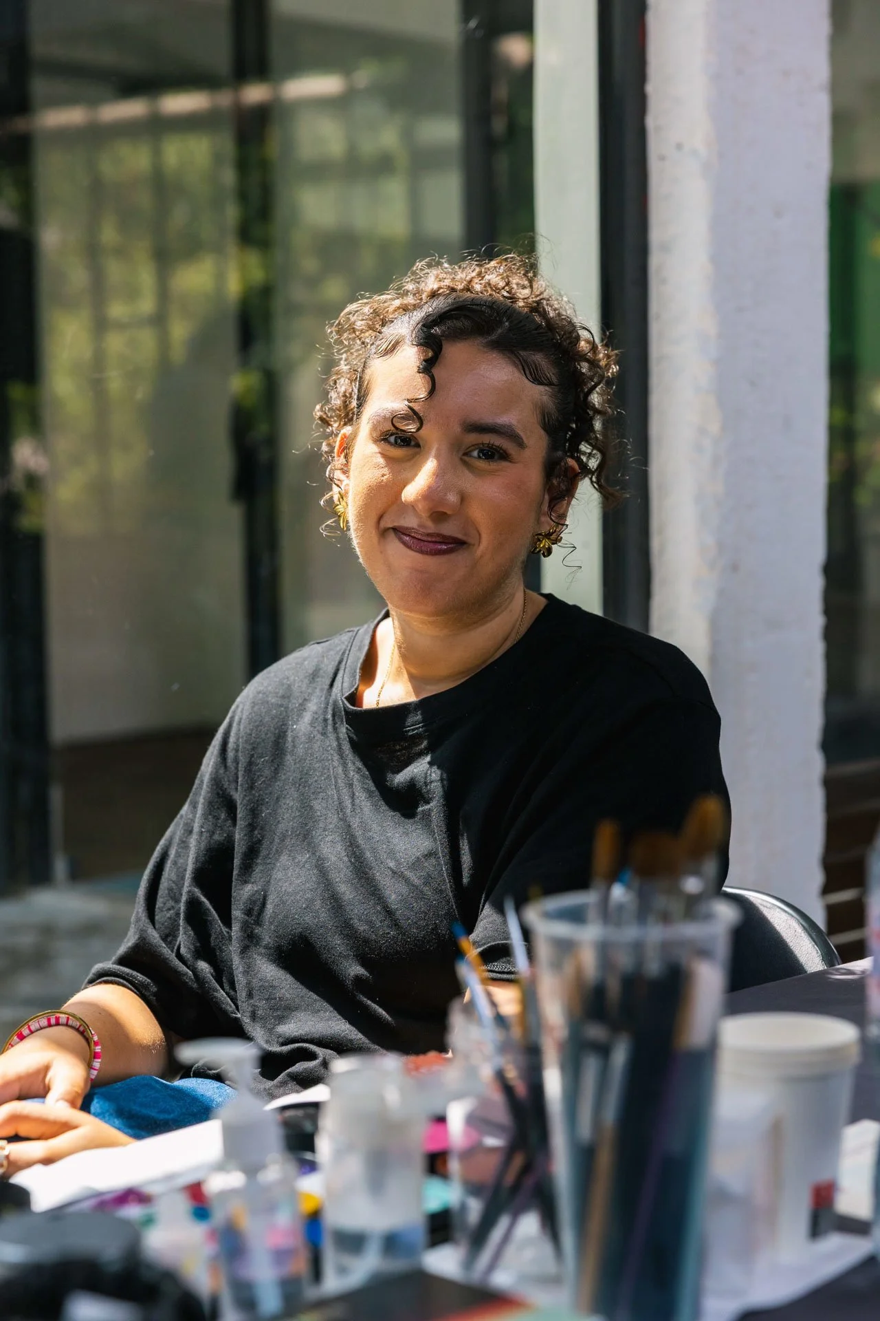 Jeune femme avec cheveux bouclés assise à une table de travail en extérieur, souriante et portant un t-shirt noir, avec divers objets comme pinceaux et pots de peinture sur la table.