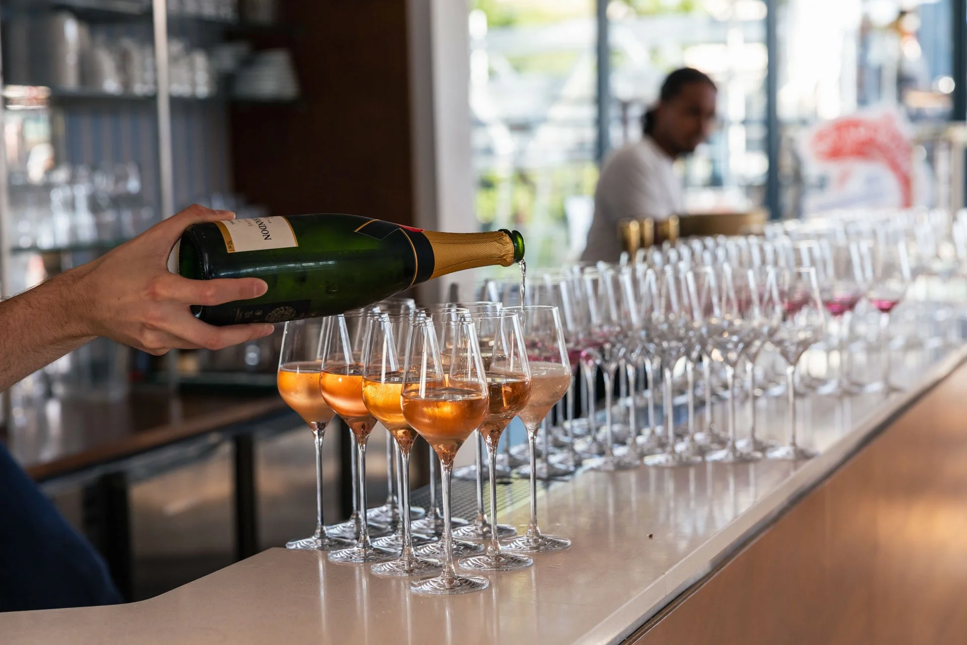 Une main verse une boisson mousseuse dans des verres à vin rose sur un bar ou une table, avec un arrière-plan flou de personnel de bar ou de restaurant.