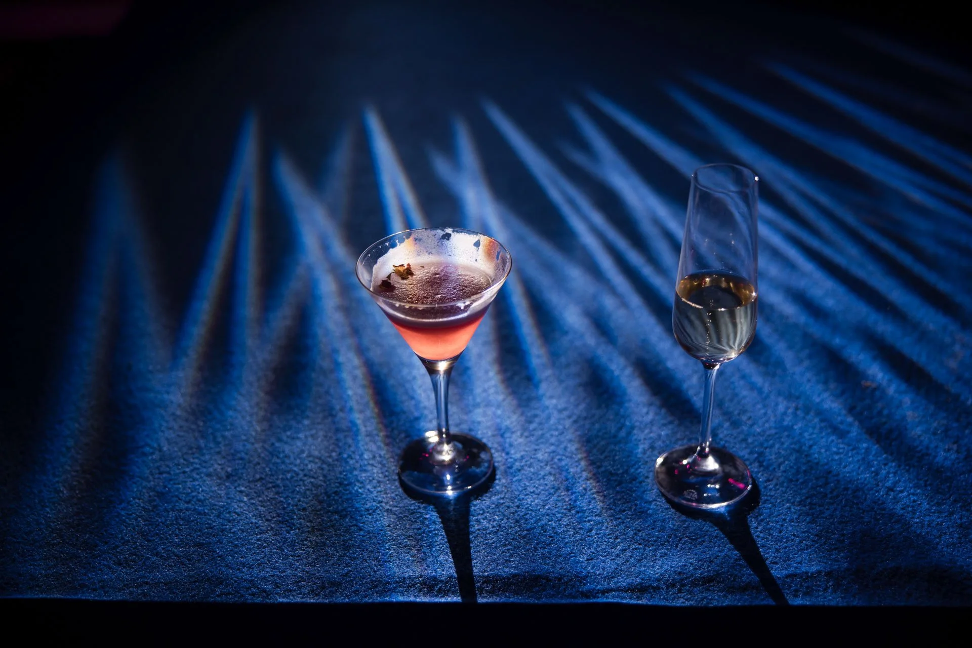 Deux verres à cocktail sur une surface sombre, éclairés par un faisceau de lumière bleue, créant des ombres longues et des reflets.