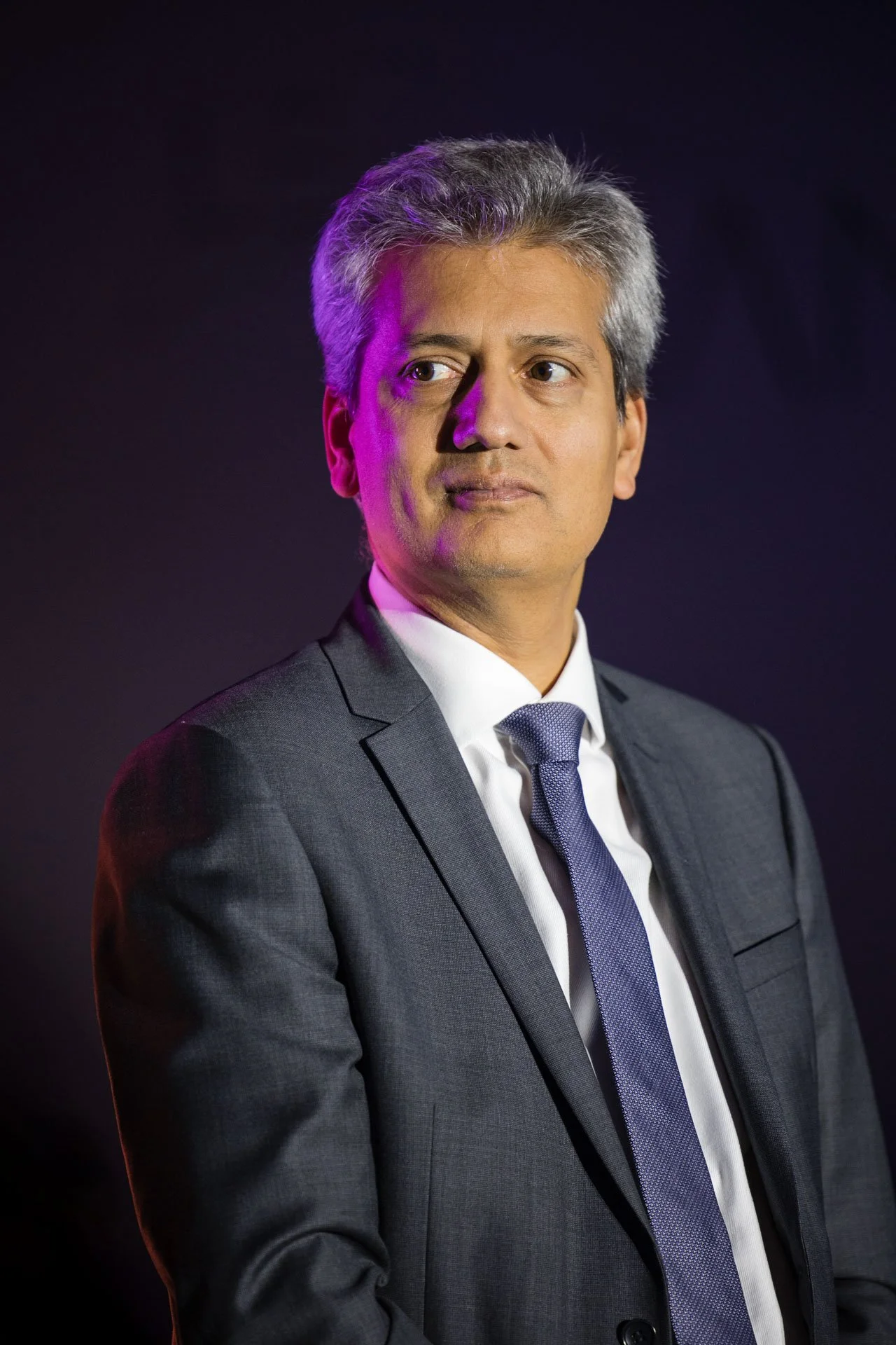 Homme en costume gris, chemise blanche et cravate, regardant à gauche, portrait en studio avec fond noir, éclairage avec une lumière violette sur le côté gauche de son visage.