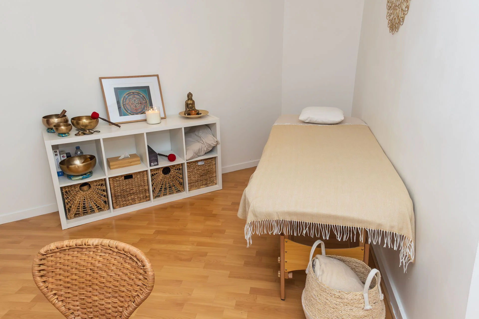 Une salle de massage avec un lit recouvert d'une couverture beige, un coussin blanc, une armoire blanche avec des objets de décoration, une statue de bouddha, une bougie, et une photo encadrée, le sol en parquet.