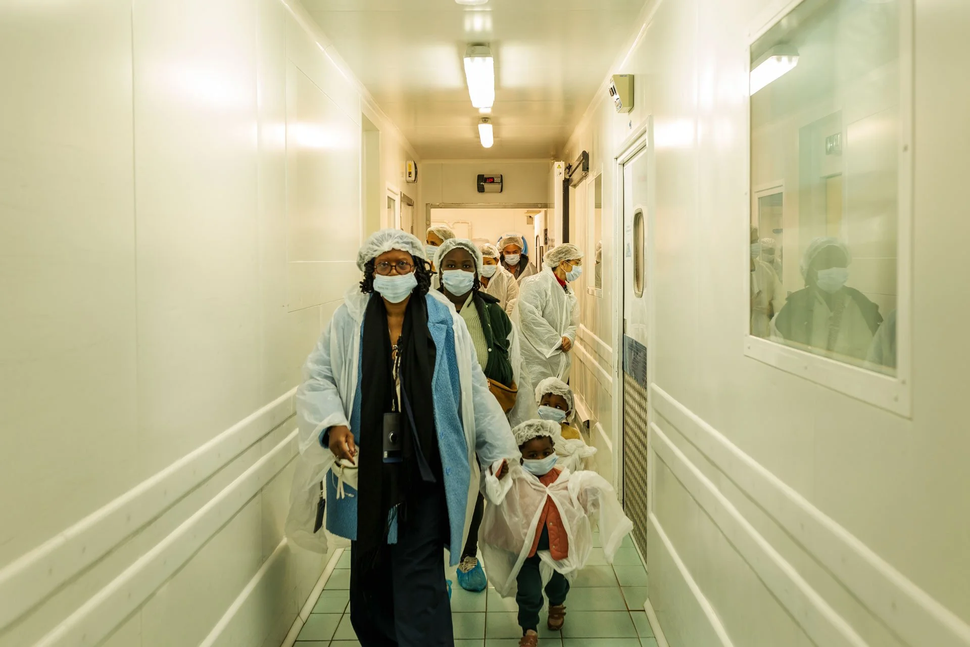 Groupe de professionnels en tenue de protection, comprenant des femmes et des enfants, marchant dans un couloir d'hôpital ou de laboratoire, tous portant des masques faciaux et des coiffes de sécurité.