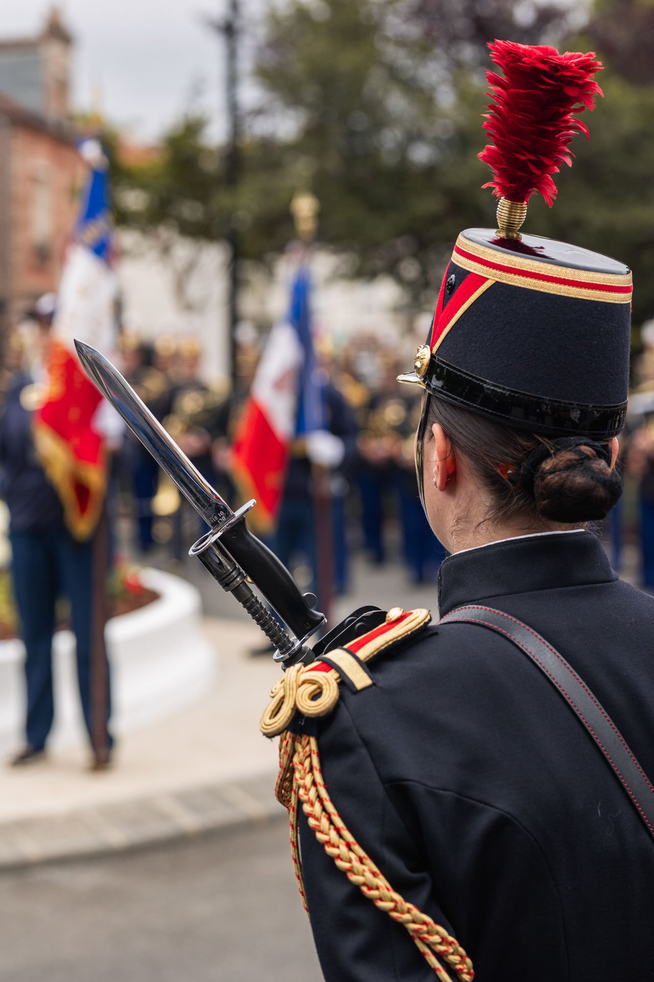 Une soldate en uniforme de cérémonie, portant un chapeau traditionnel avec une plume rouge, et tenant une épée, lors d'une cérémonie officielle ou une parade avec un groupe de soldats en arrière-plan portant des drapeaux français.
