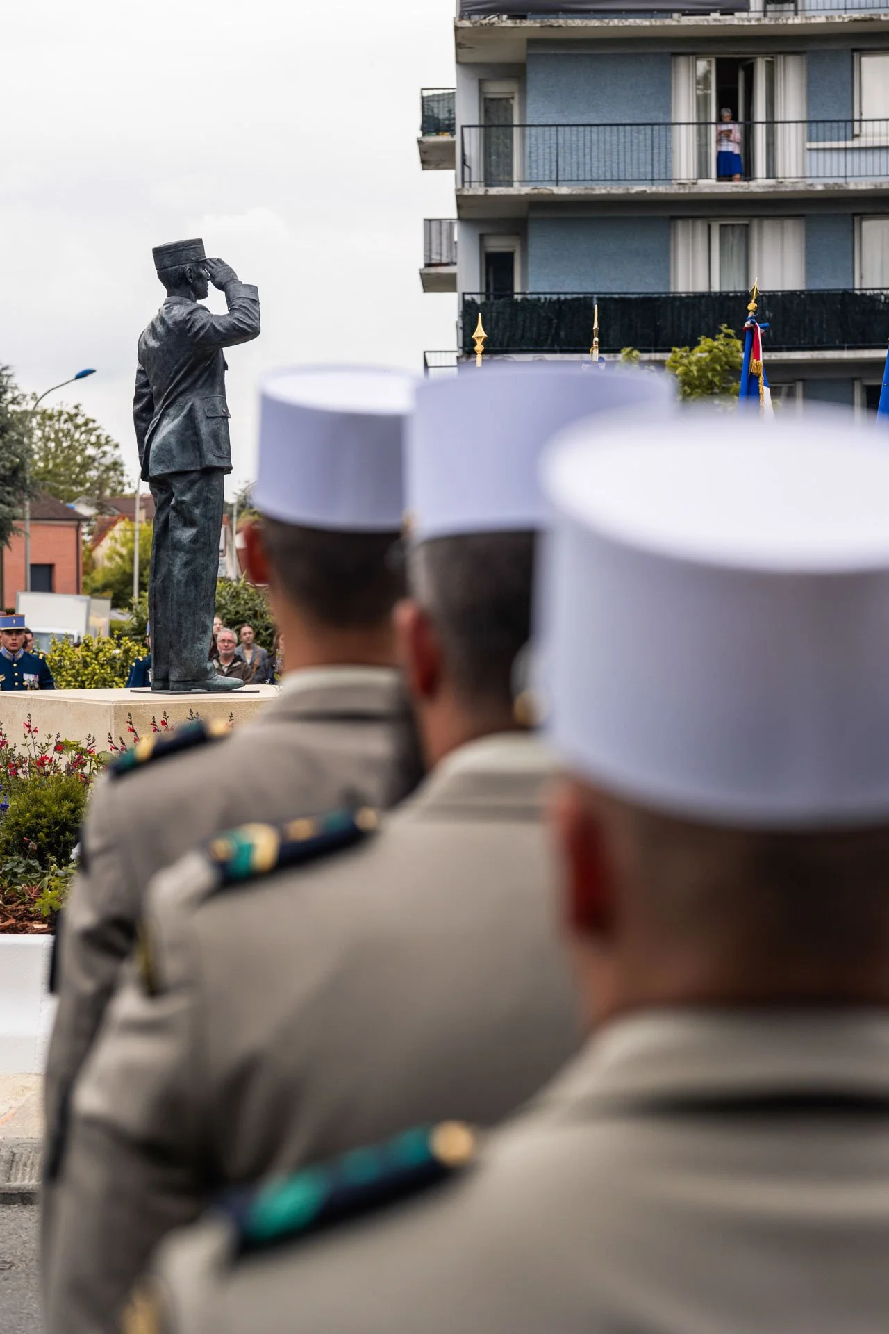 Statue d'un homme saluant lors d'une cérémonie officielle, avec des militaires en uniforme en première ligne et un bâtiment résidentiel en arrière-plan.