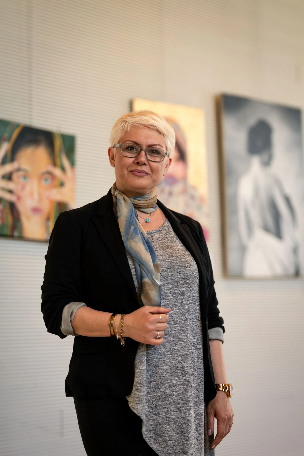 Une femme aux cheveux blonds courts et lunettes, portant un blazer noir, un haut gris, une écharpe colorée, des bracelets et une montre dorée, se tenant devant des œuvres d'art abstraites dans une galerie.