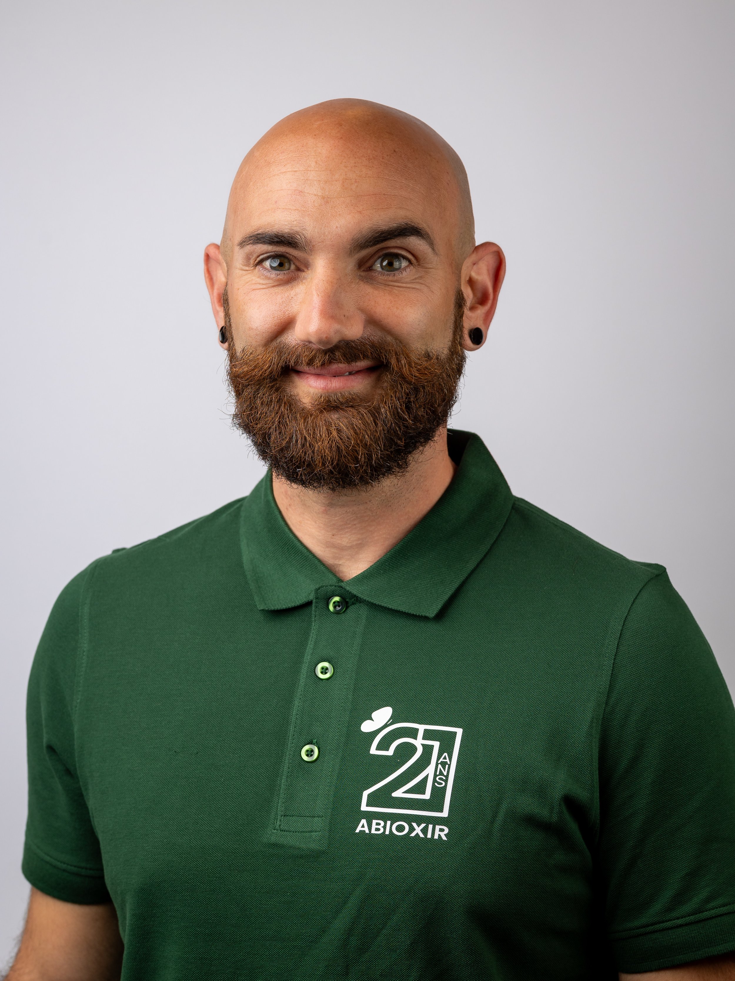 Un homme chauve avec une barbe brune, portant un polo vert avec un logo blanc. Il sourit légèrement.