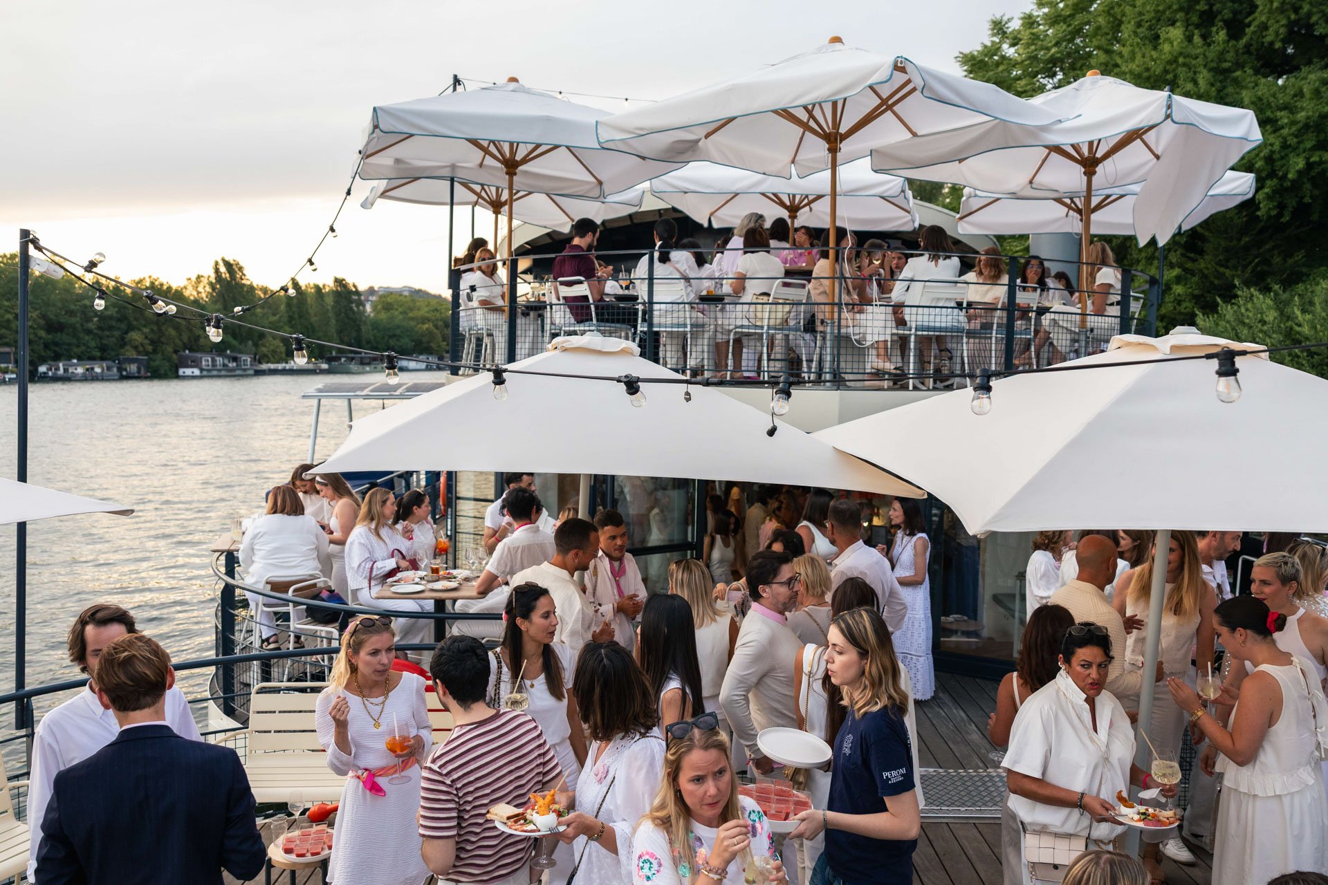 Une terrasse en extérieur au bord d'une rivière, remplie de personnes élégantes en tenue blanche ou claire, dégustant des plats et des boissons, avec des parasols et des guirlandes lumineuses.
