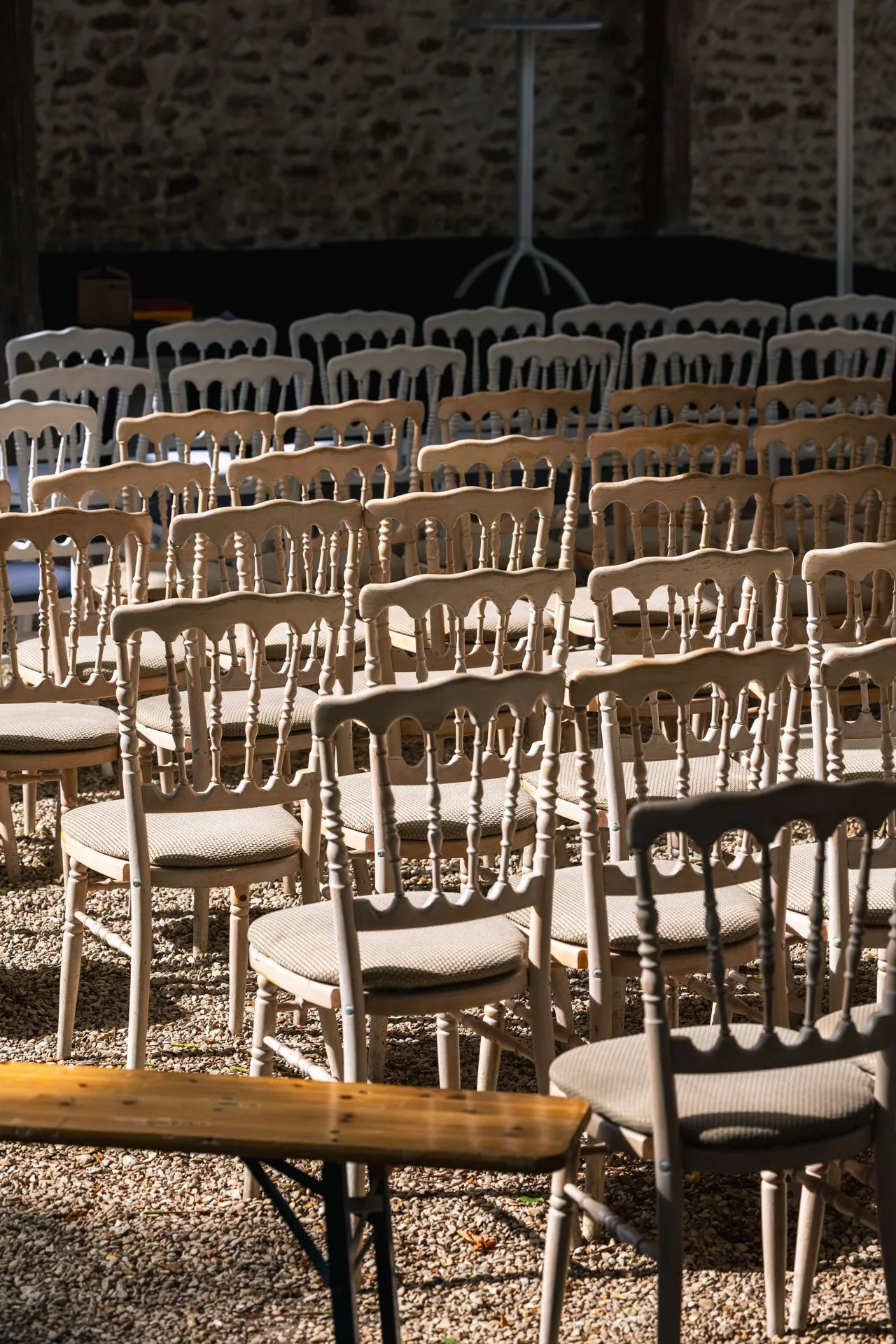 rangée de chaises en bois et tissu beige au sol en gravier, dans un espace intérieur avec mur en pierre et éclairage