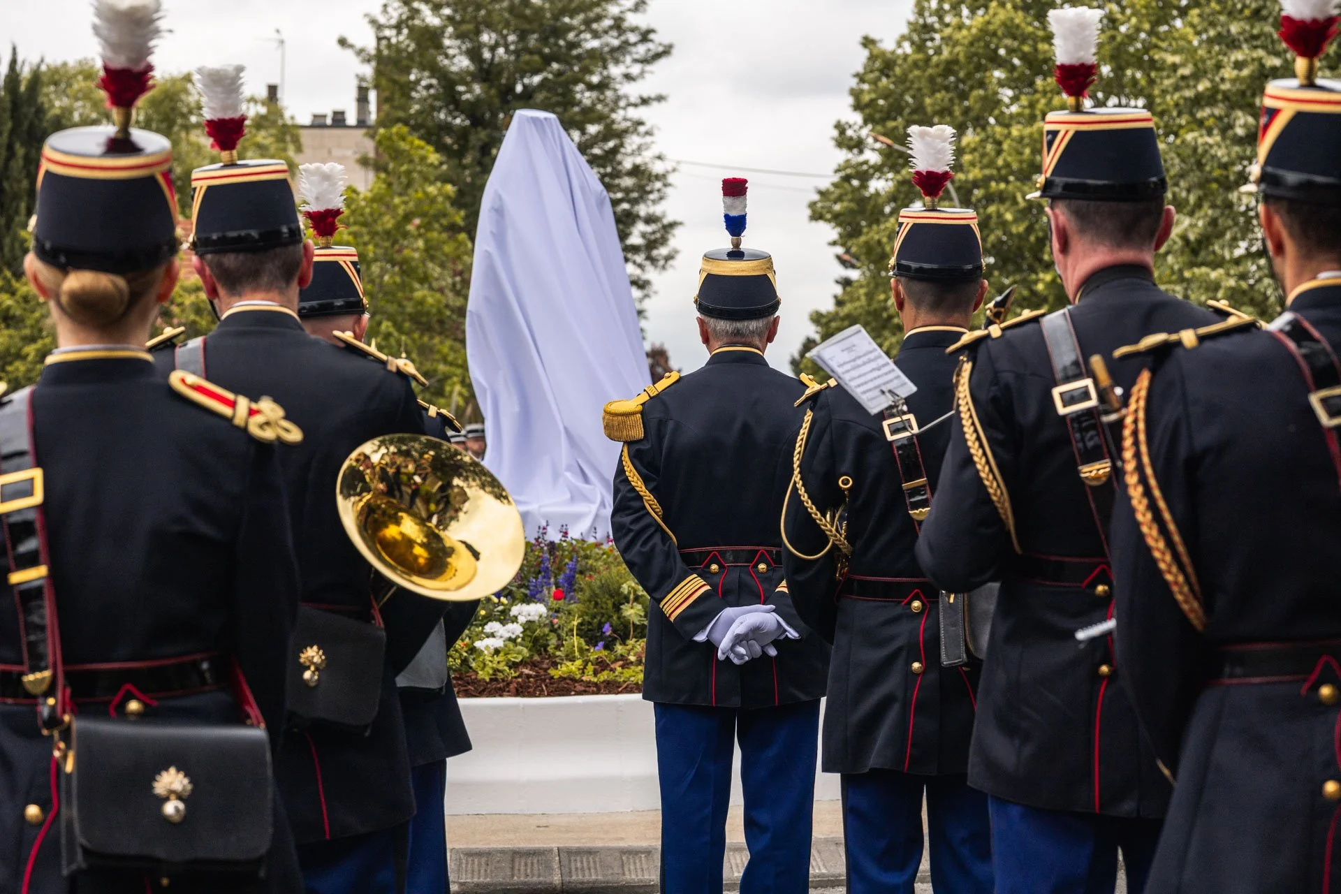 Groupe de musiciens en uniformes militaires avec des chapeaux à plumes, rassemblés lors d'une cérémonie commémorative en plein air, avec une structure couverte blanc en arrière-plan.