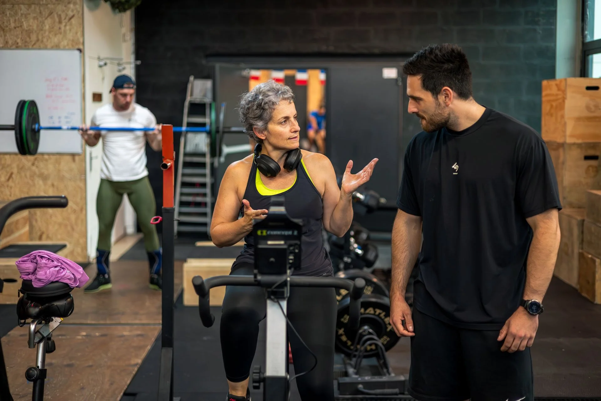 Une femme et un homme parlent dans une salle de sport. La femme porte un casque autour du cou et une tenue de sport noire avec du jaune. L'homme porte un t-shirt noir. En arrière-plan, un homme en t-shirt blanc soulève une barre de musculation.