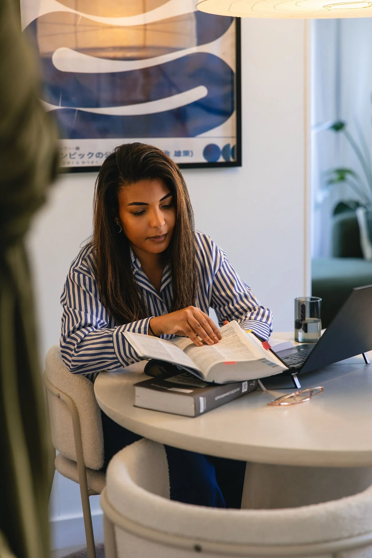 Une femme assise à une table, lisant un livre avec un ordinateur portable ouvert devant elle, dans un environnement intérieur.