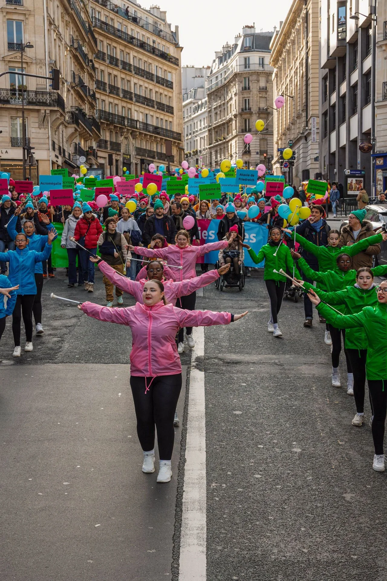 Un groupe de personnes participe à une marche ou un rassemblement dans une avenue de Paris, avec des bâtiments haussmanniens en arrière-plan. Certaines personnes portent des vêtements colorés, et beaucoup tiennent des ballons et des pancartes avec de