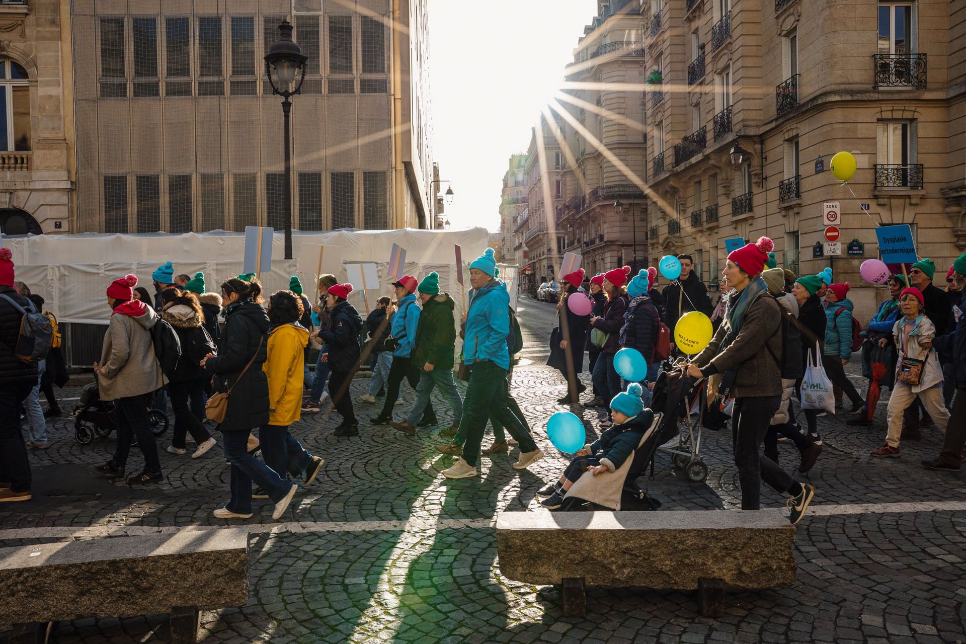 Une foule de personnes marchent dans une rue de ville ensoleillée, portant des bonnets colorés, certains tenant des ballons et des pancartes, avec un bâtiment ancien à droite et un immeuble moderne au fond.