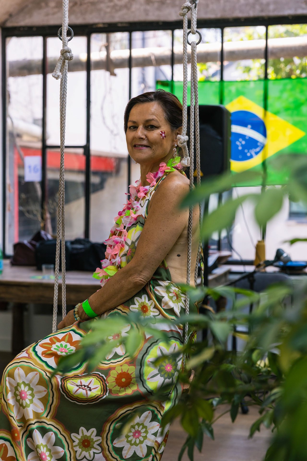 Une femme souriante portant une robe colorée à motifs floraux, une parure de fleurs et un bracelet vert, assise sur une balançoire en intérieur, avec un drapeau du Brésil en arrière-plan.