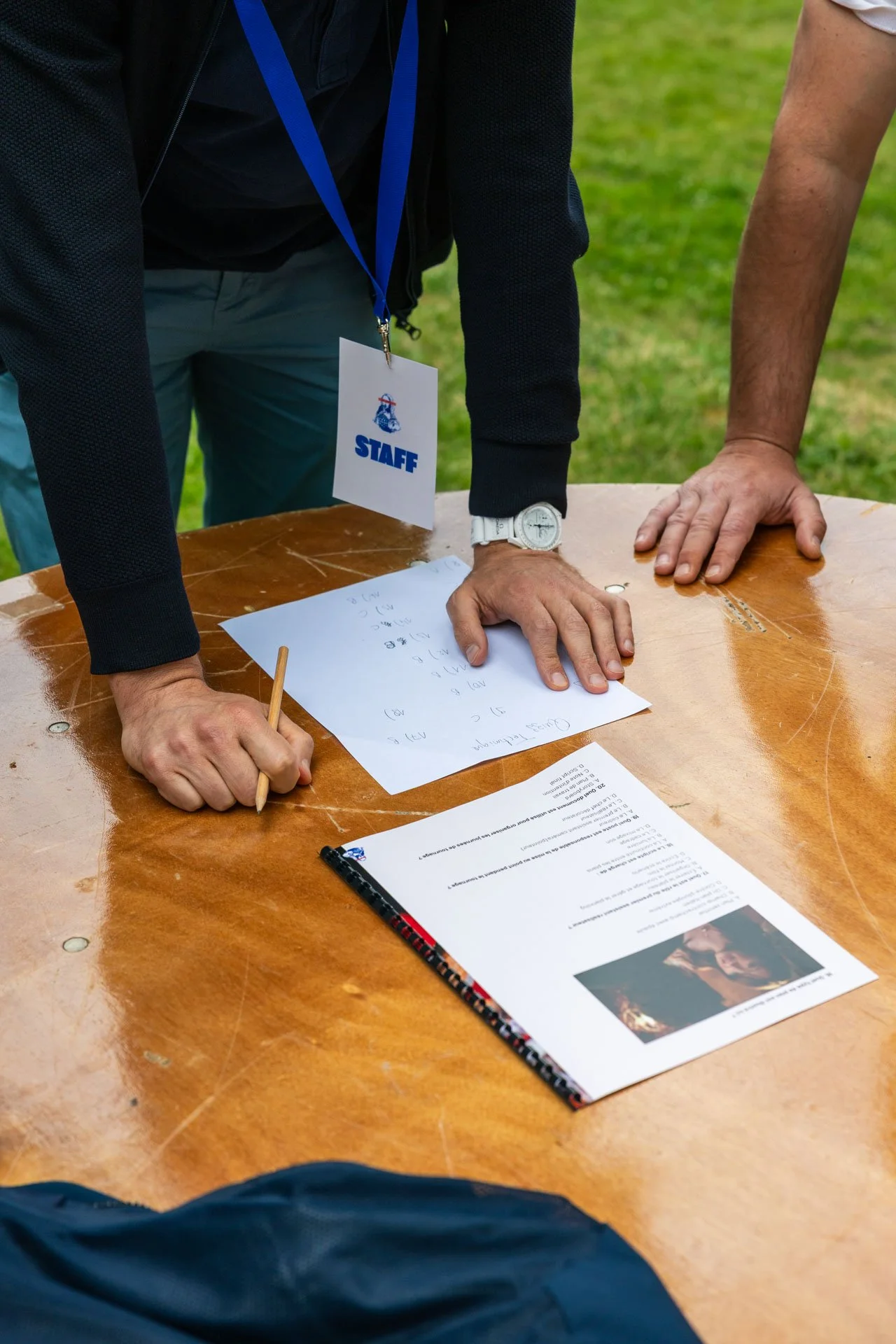 Groupe de personnes debout autour d'une table en bois lors d'une réunion ou d'un événement extérieur. L'une d'elles porte un badge avec l'inscription 'STAFF'. Il y a des documents et un carnet sur la table.