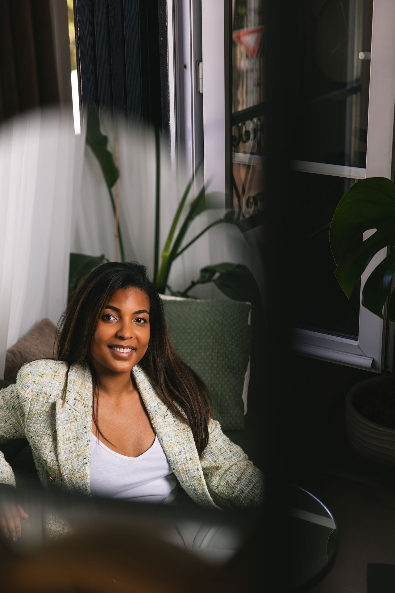 Une femme souriante avec des cheveux longs bruns, portant une veste beige à carreaux et une chemise blanche, assise dans un espace intérieur avec des plantes vertes et un canapé en arrière-plan.