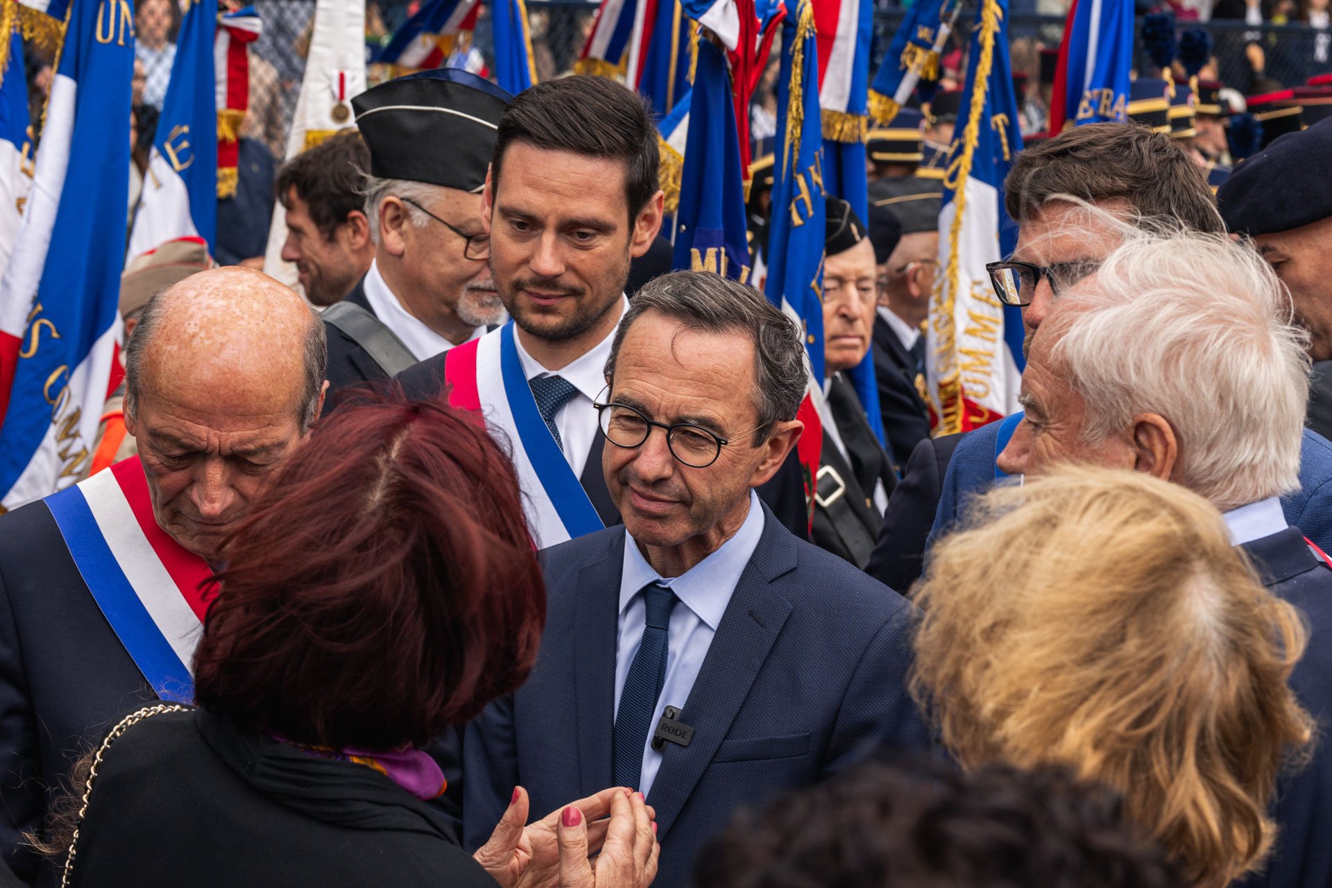 Groupe de personnes lors d'une cérémonie officielle, avec des drapeaux français en arrière-plan, certaines personnes portent des écharpes tricolores.