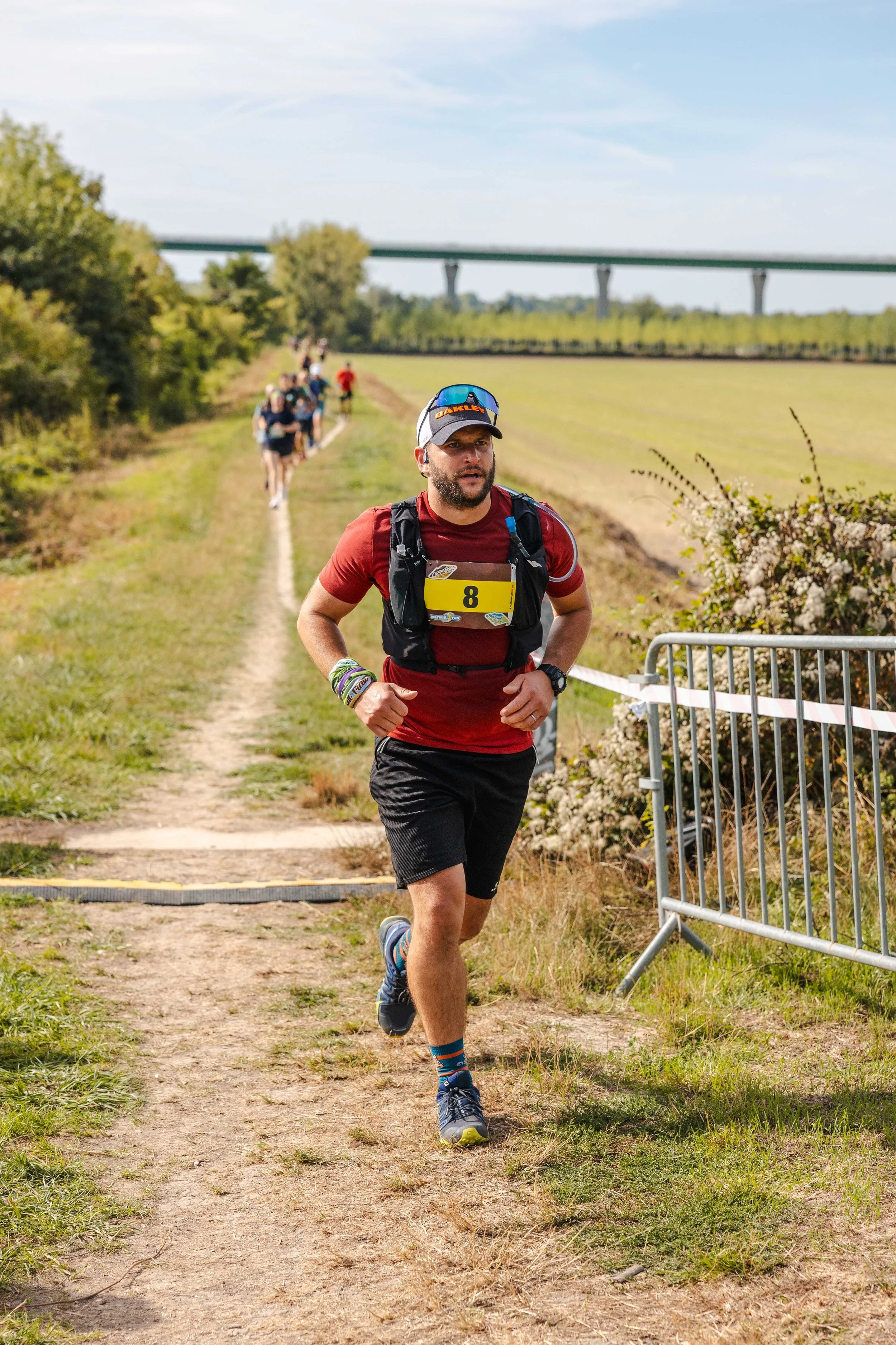 Homme courant lors d'une course en plein air, portant un numéro 8, une casquette et un équipement de course, avec d'autres coureurs en arrière-plan sur un sentier en campagne.
