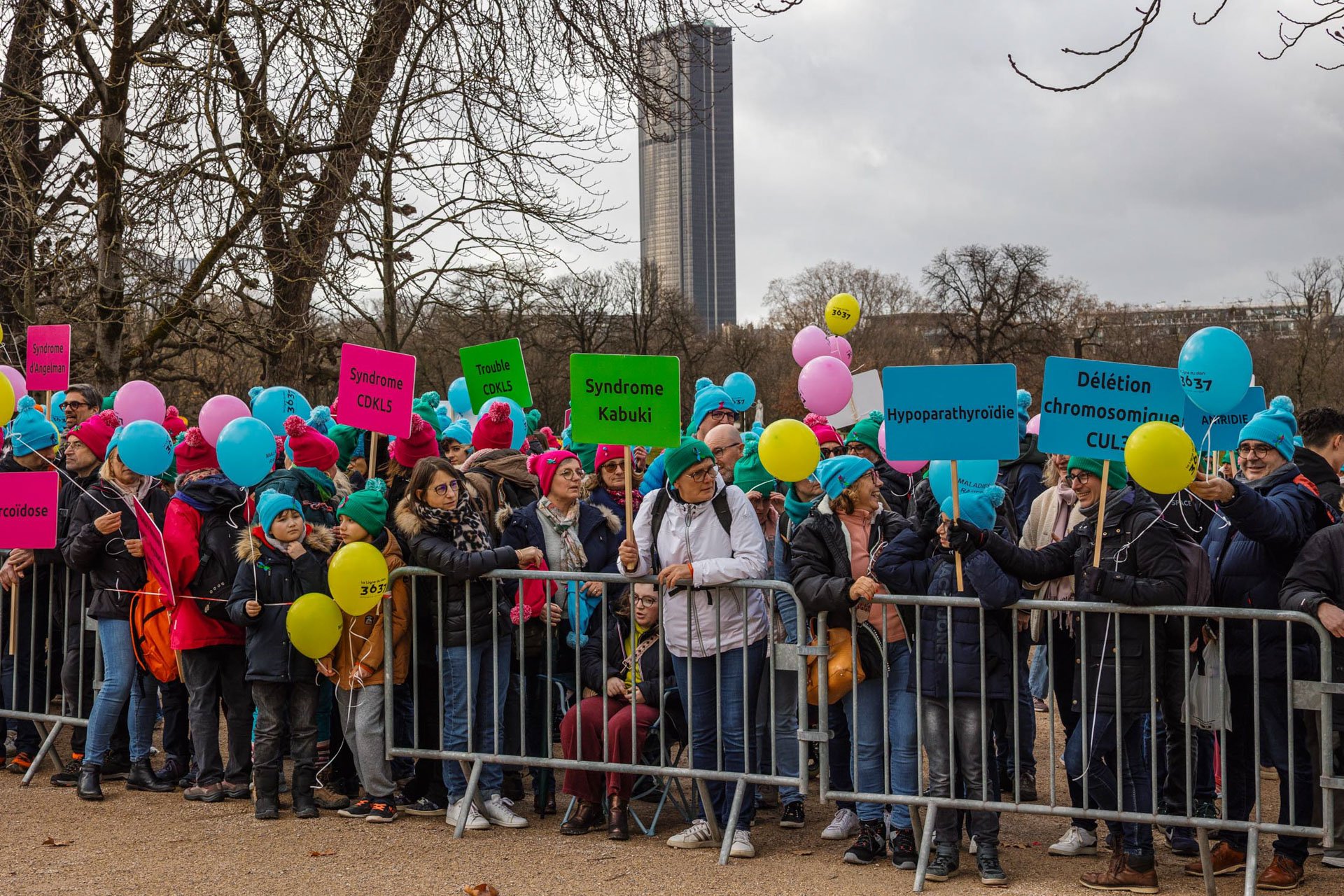Groupe de personnes participant à une marche ou à une manifestation, tenant des ballons colorés et des pancartes avec des noms de syndromes médicaux, dans un parc avec des arbres nus et un bâtiment en arrière-plan.