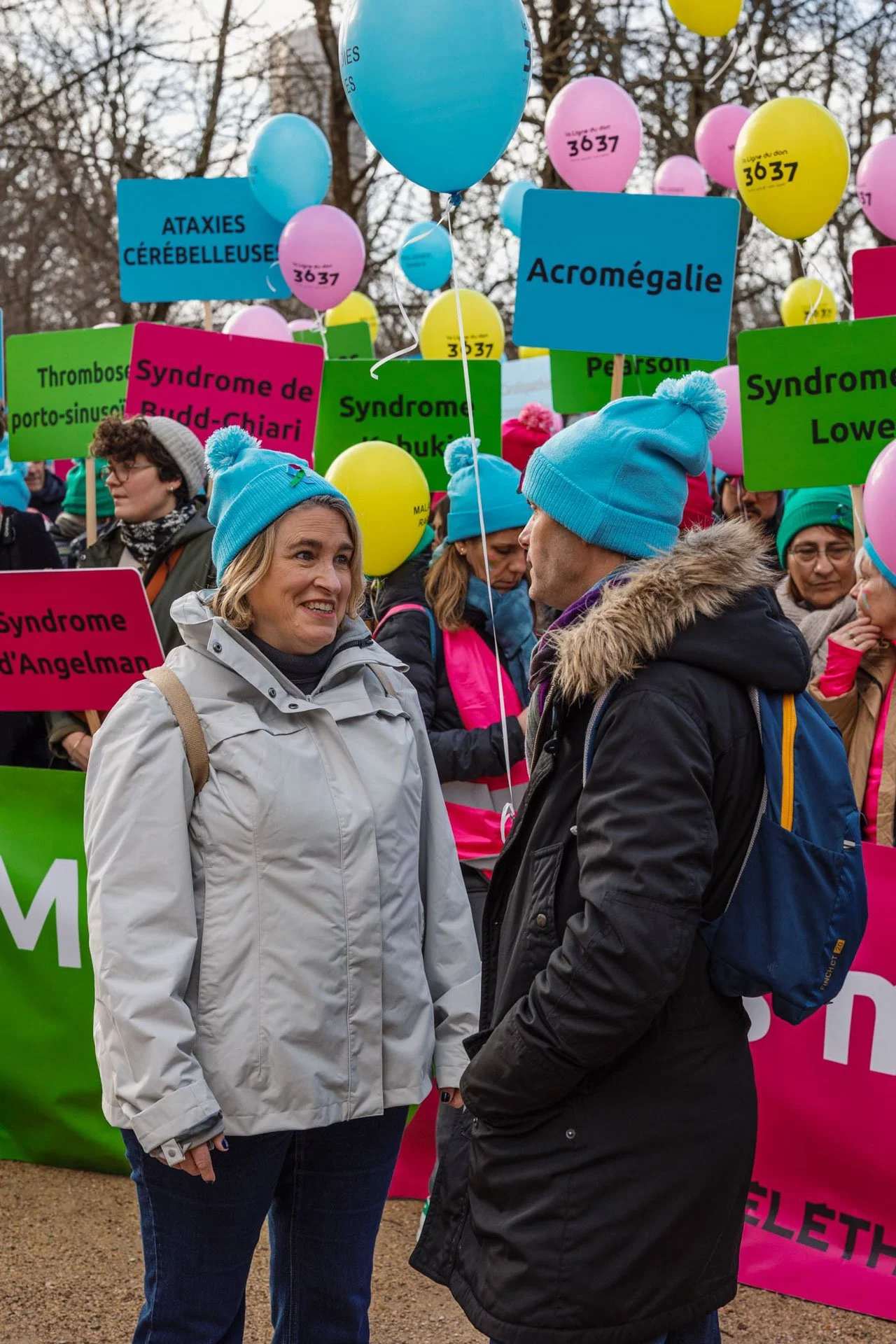 Deux personnes discutent lors d'une manifestation ou rassemblement, avec des panneaux et ballons colorés portant des noms de syndromes et de troubles, comme syndrome de Down, syndrome de Budd-Chiari, acromégalie, aperture, et autres.
