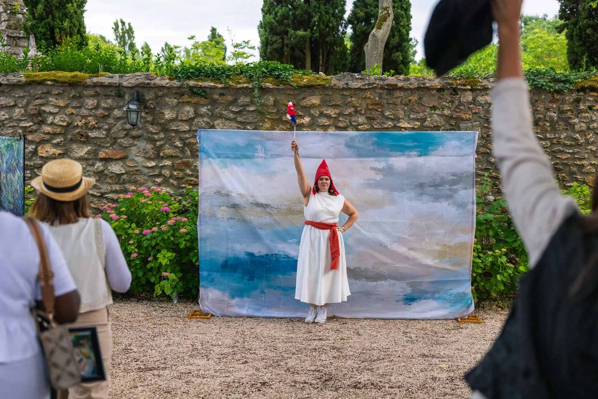 Une femme déguisée en personnage historique ou mythologique pose devant un fond peint représentant un ciel et des nuages, lors d'une performance ou d'une séance de théâtre en plein air, avec des spectateurs regardant.
