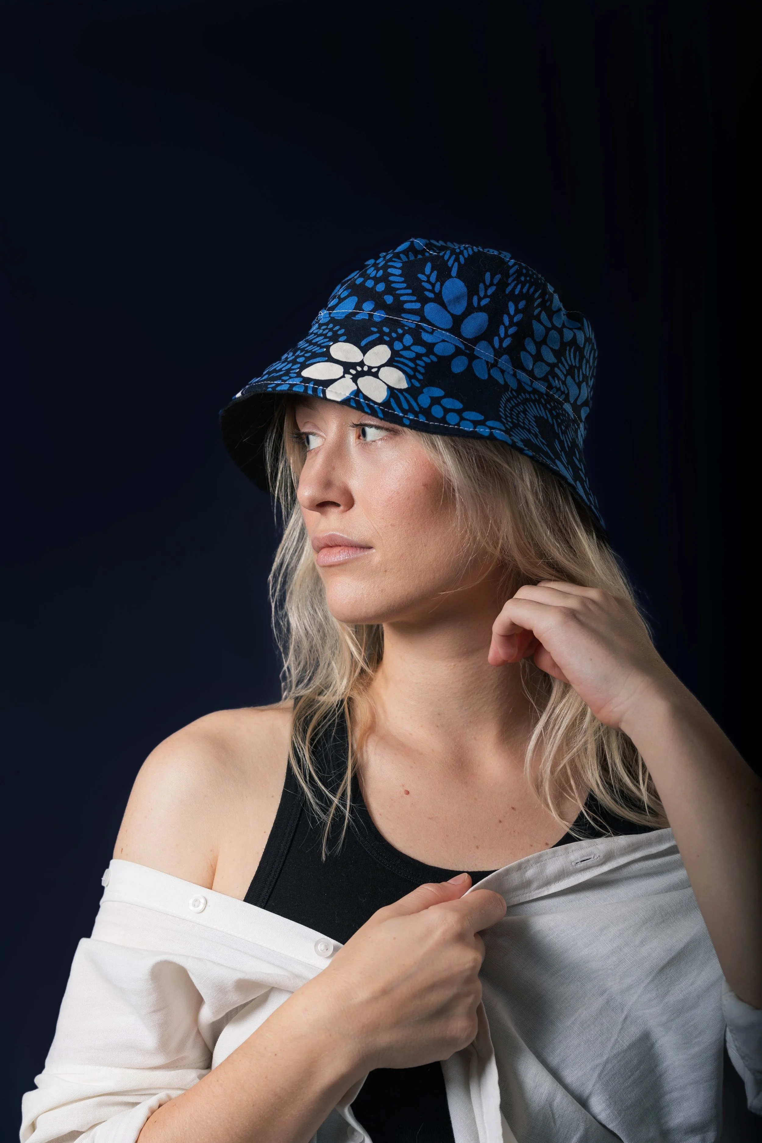 Une femme avec des cheveux blonds portant un chapeau de style bucket avec un motif floral bleu et blanc, regardant vers la gauche, contre un fond noir