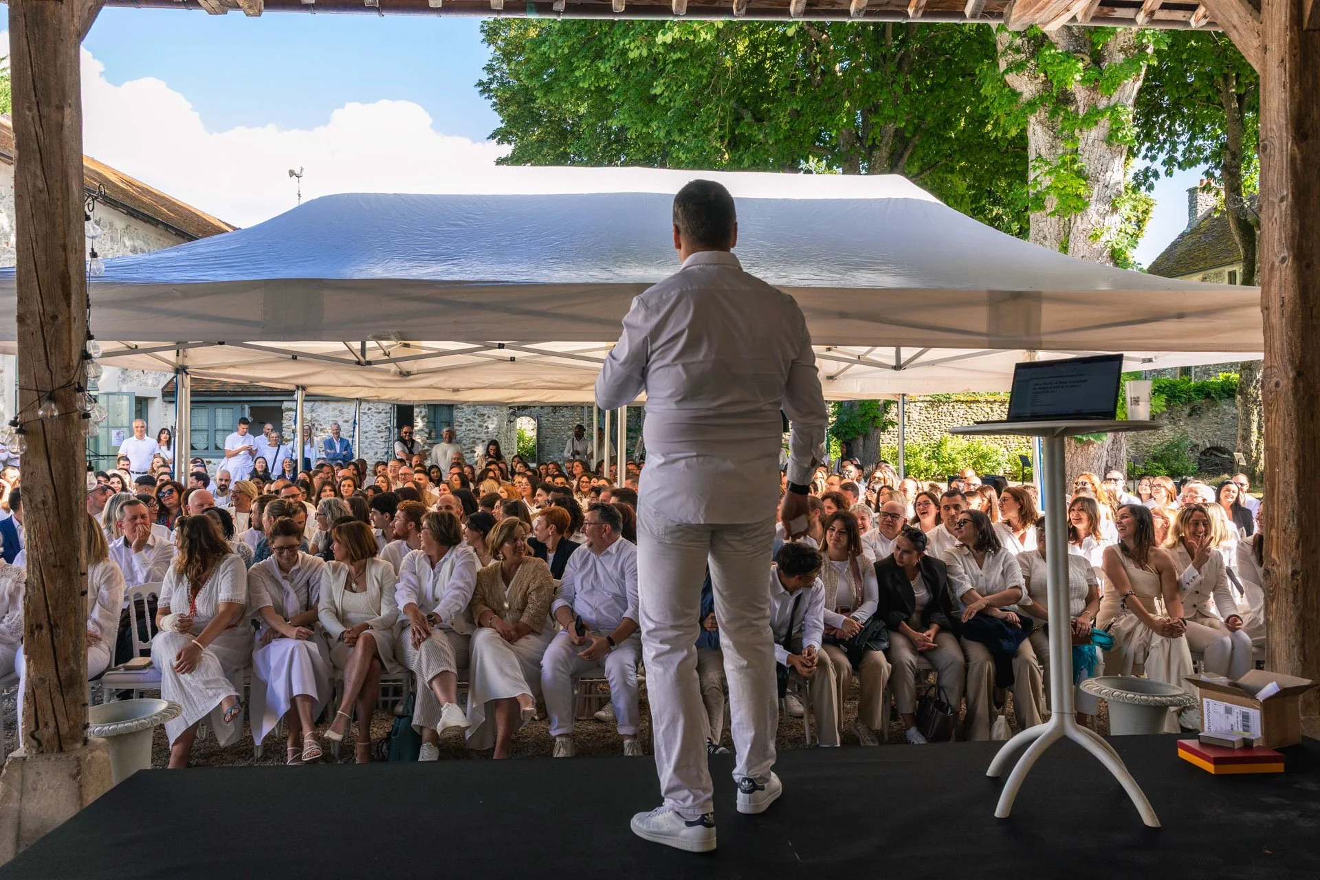 Un homme debout sur scène face à une grande audience lors d'un événement en plein air, sous une tente, avec des arbres en arrière-plan.