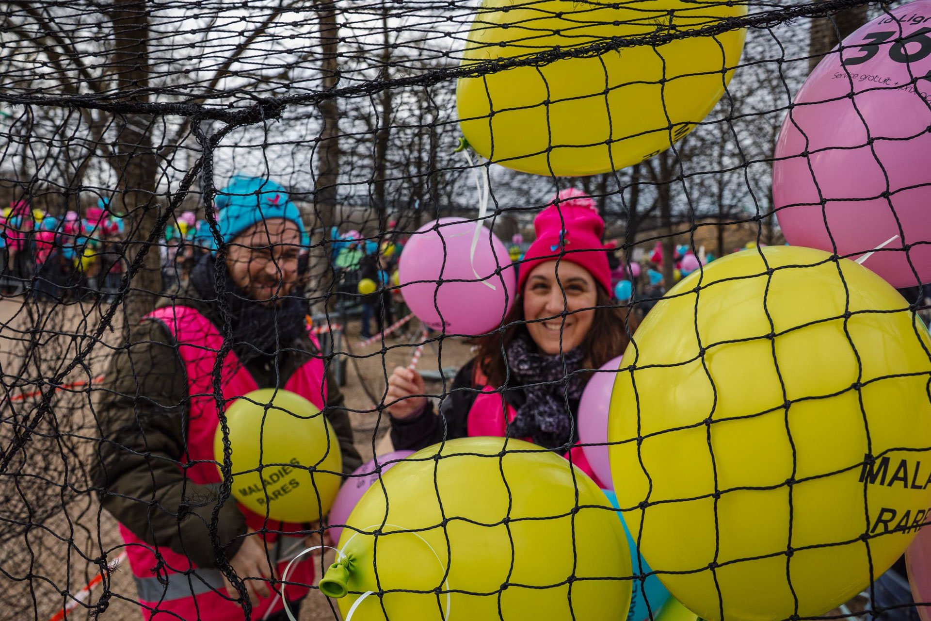 Deux femmes souriantes derrière une clôture en filet avec des ballons jaunes et roses dans une fête en plein air.