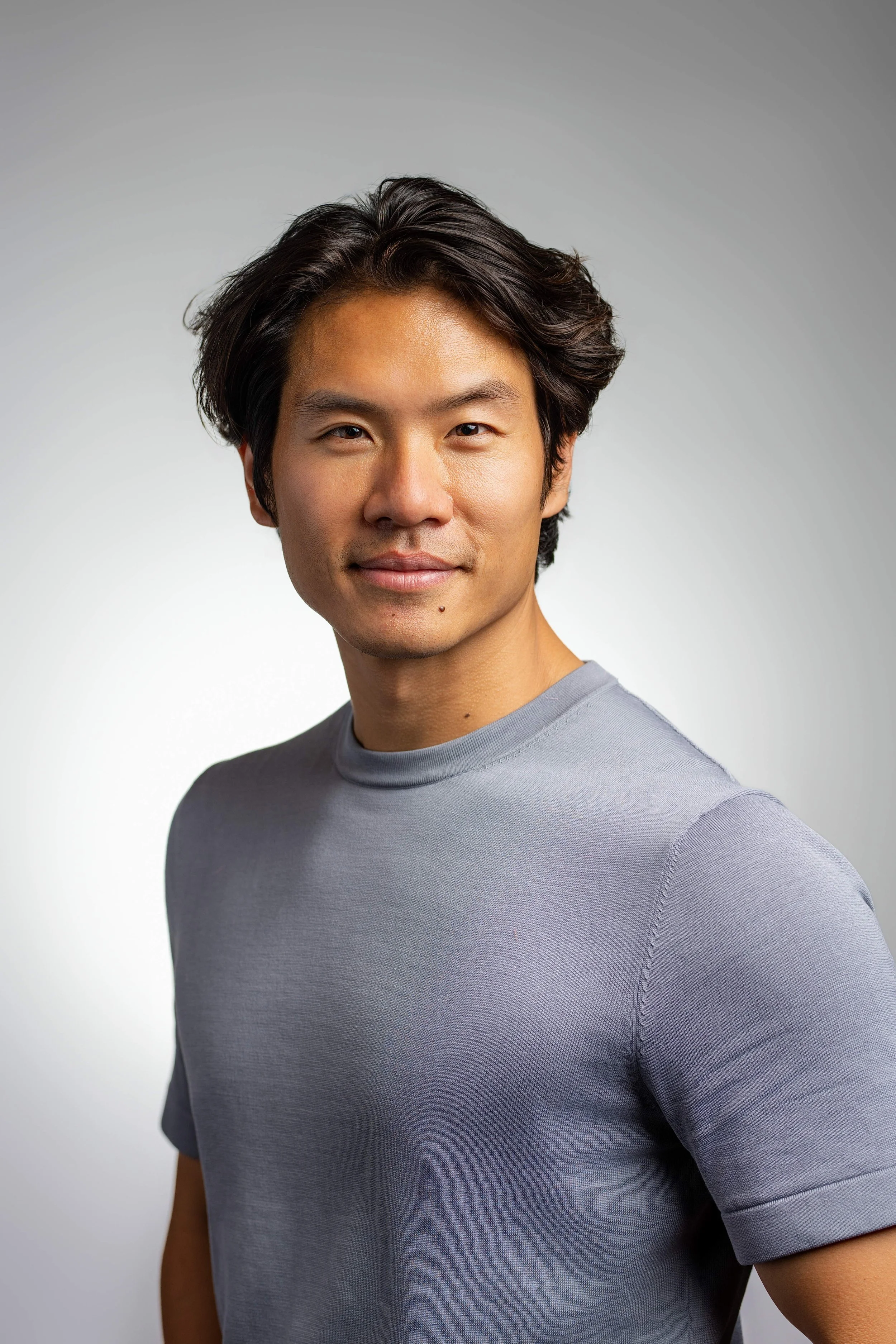 Portrait d'un jeune homme asiatique avec des cheveux noirs ondulés, portant un t-shirt gris clair, souriant légèrement, avec un fond gris clair