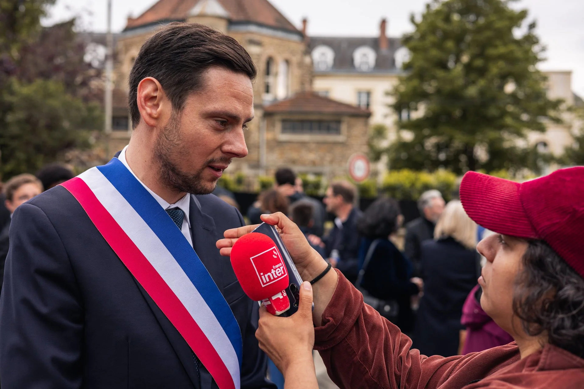 Un homme portant une veste sombre avec un ruban tricolore (bleu, blanc, rouge) reçoit une interview d'une femme portant un chapeau rouge, lors d'une cérémonie ou événement public en extérieur, avec des bâtiments anciens et des arbres en arrière-plan.