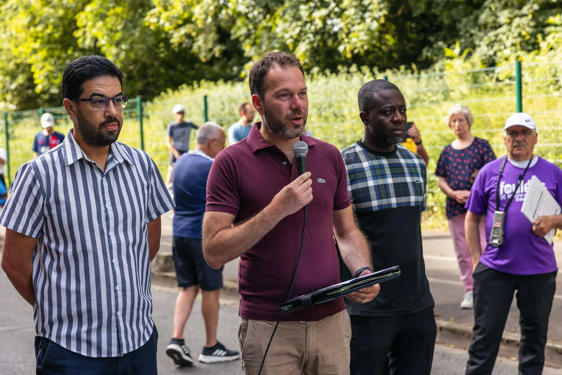 Un homme parle dans un micro lors d'une manifestation ou événement public, entouré de plusieurs personnes dans un cadre extérieur avec des arbustes en arrière-plan.