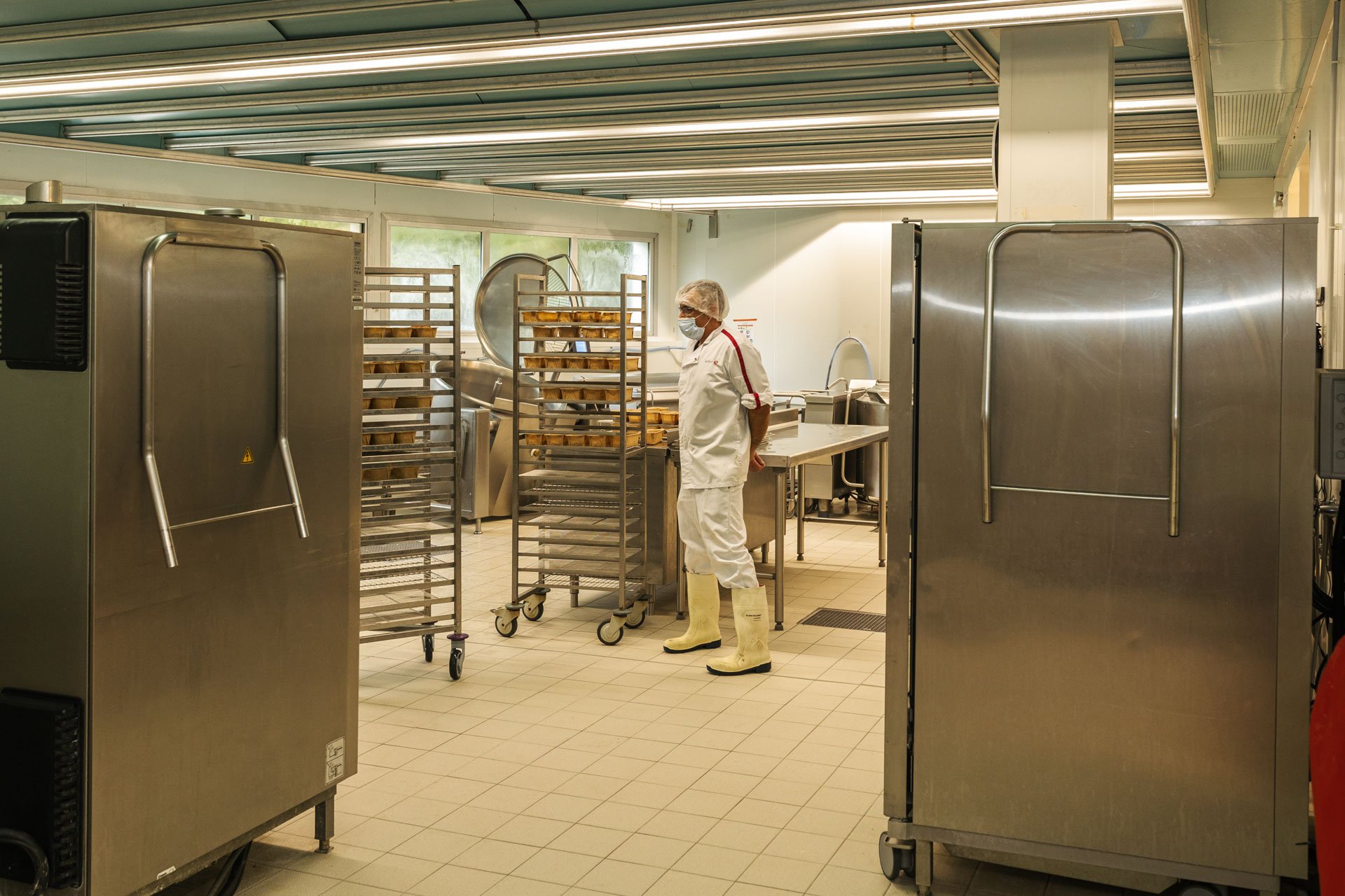 Une cuisine industrielle avec un personnel en tenue de protection, portant des bottes en caoutchouc et un masque, entouré de rayonnages en acier et de grandes machines de cuisson ou de stockage.