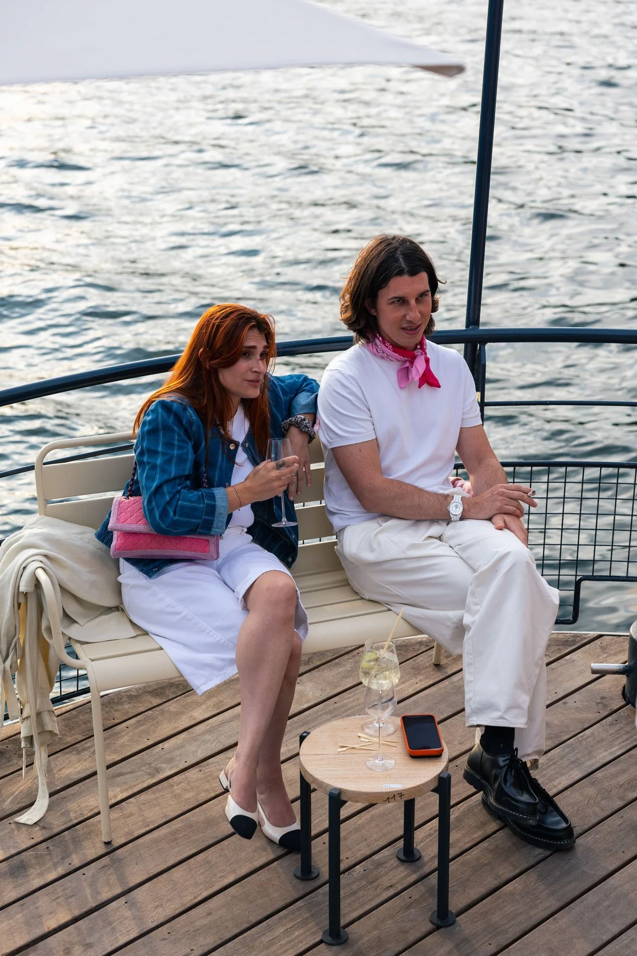 Deux personnes assises sur un banc en bateau avec vue sur l'eau. Une femme avec une robe blanche, une veste en jean et des chaussures blanches à talons, tenant un verre. Un homme avec un t-shirt blanc, un bandana rose, un bracelet blanc, pantalon bla