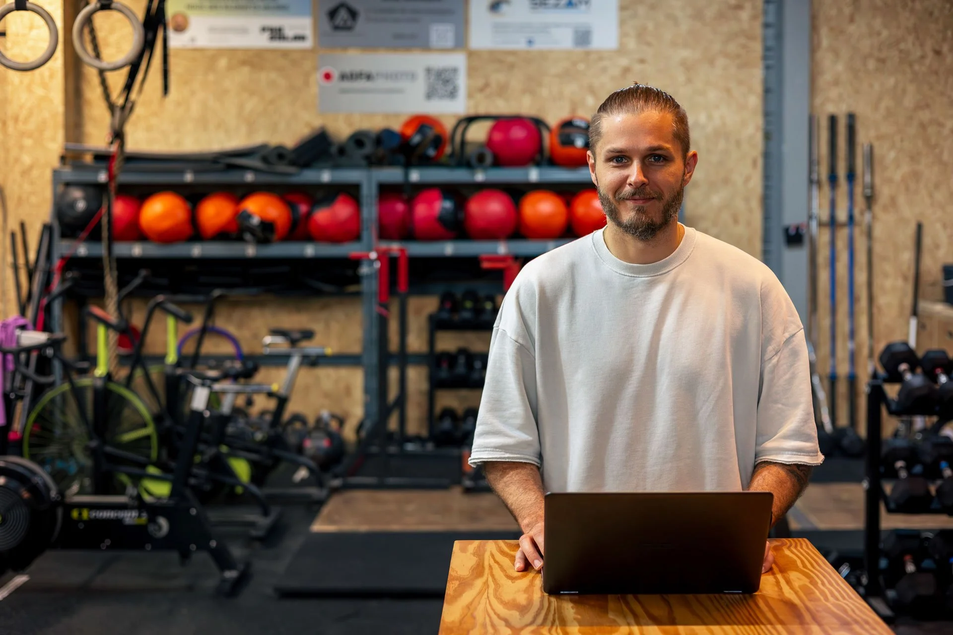 Un homme souriant regarde l'appareil photo dans une salle de sport, en arrière-plan il y a des équipements de fitness et des ballons de sport rouges et oranges sur une étagère.
