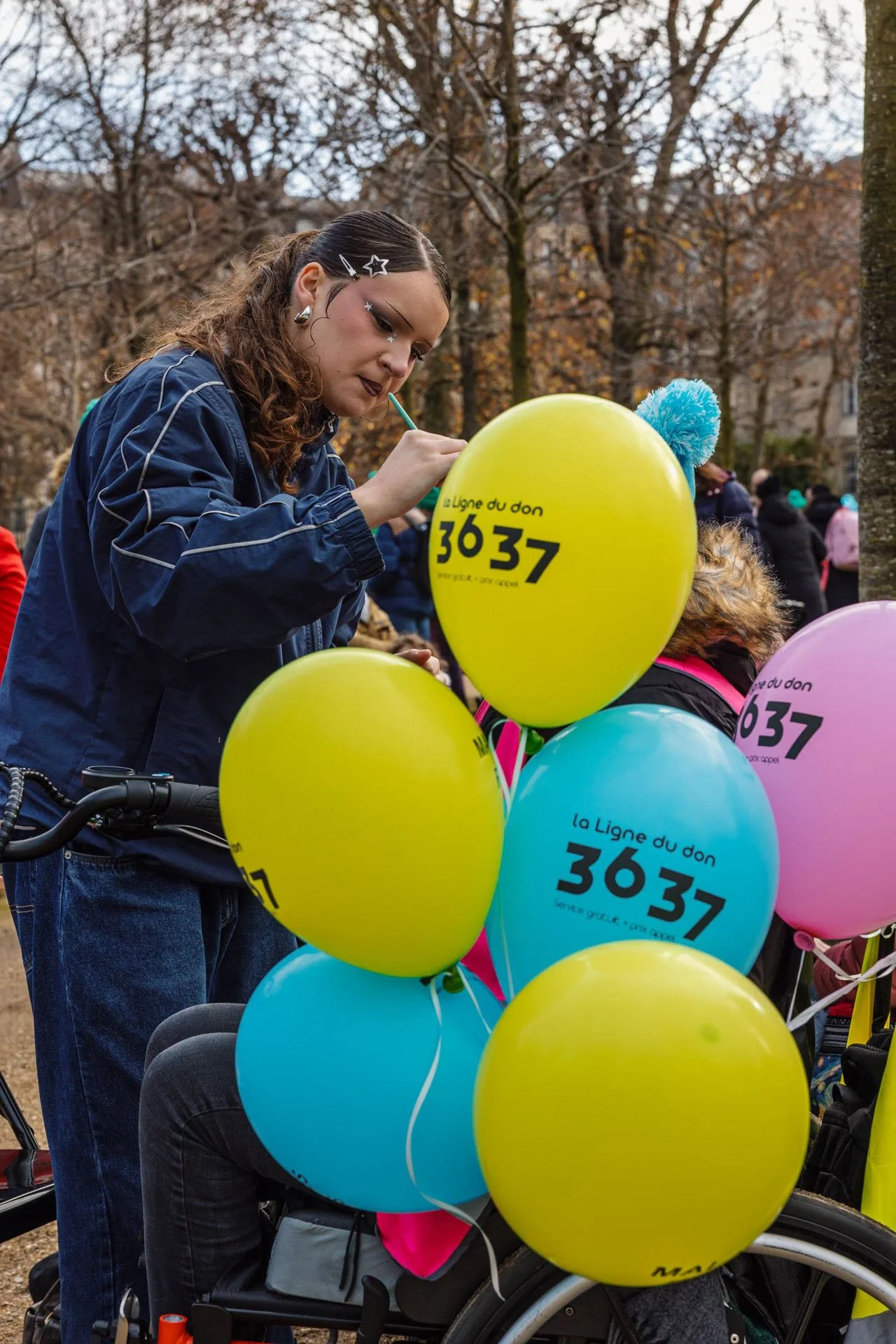Une femme avec des cheveux bouclés attachés, portant une veste bleue, qui écrit sur des ballons jaunes, bleus et roses portant le message "la Ligne du don 3637" lors d'un événement en plein air avec plusieurs personnes visibles en arrière-plan, dans 