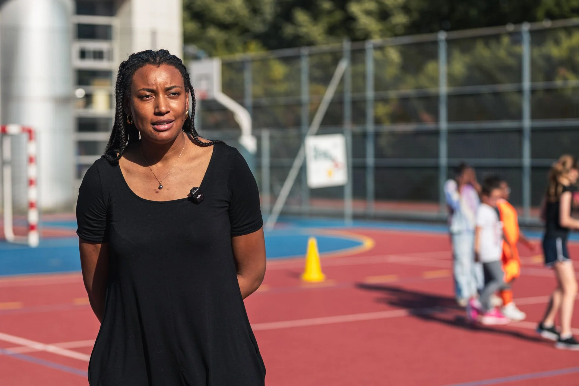 Une femme en posture de témoignage ou de discours, debout sur un terrain de sport en plein air, avec des enfants en arrière-plan dans un espace d'entraînement ou de récréation.
