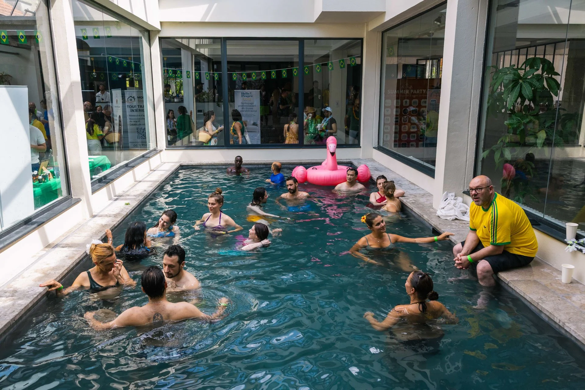 Piscine intérieure avec plusieurs personnes nageant, discutant, et se détendant, décorée avec des drapeaux brésiliens, un flamant rose gonflable, et un personnel à côté de la piscine