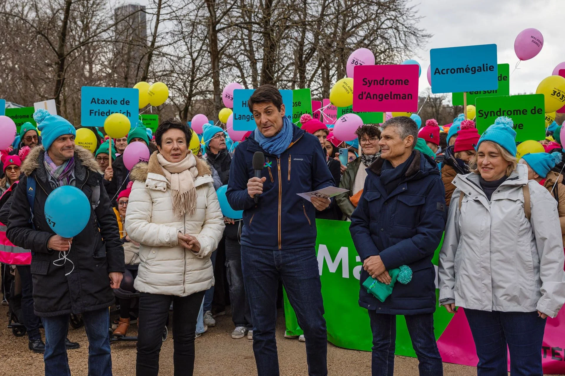 Groupe de personnes lors d'une marche ou manifestation, tenant des ballons colorés et des panneaux avec des noms de syndromes ou maladies rares, en plein air avec des arbres en arrière-plan.