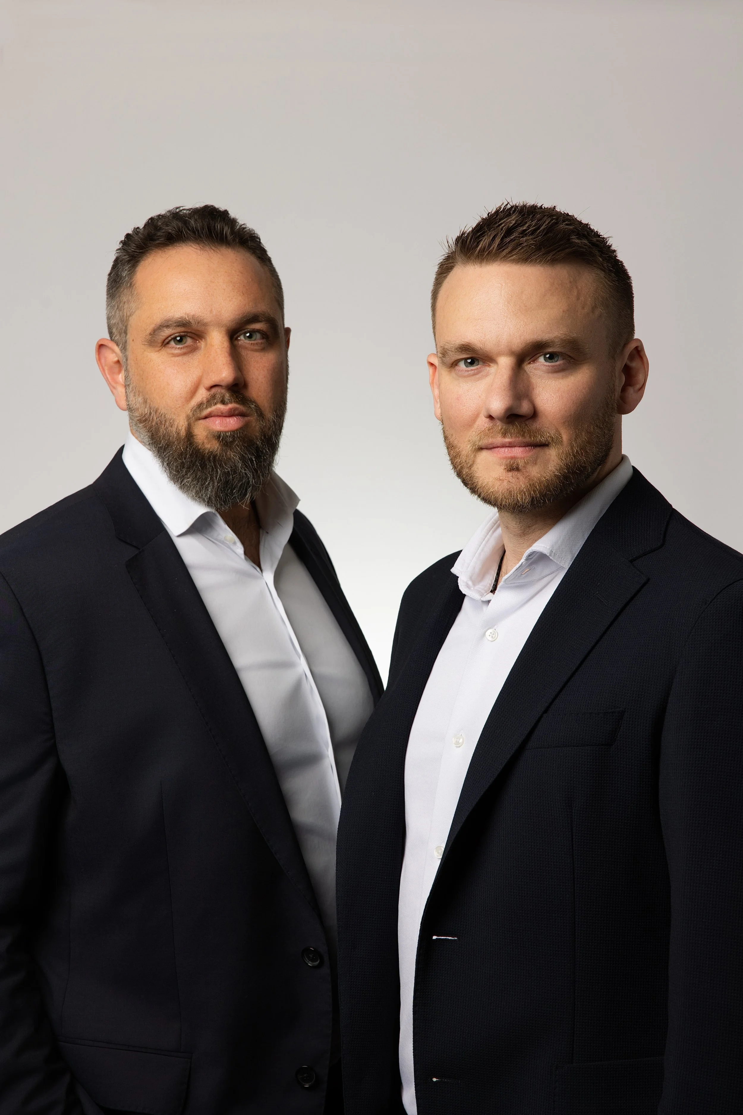 Deux hommes en costume noir et chemises blanches posent devant un fond neutre.