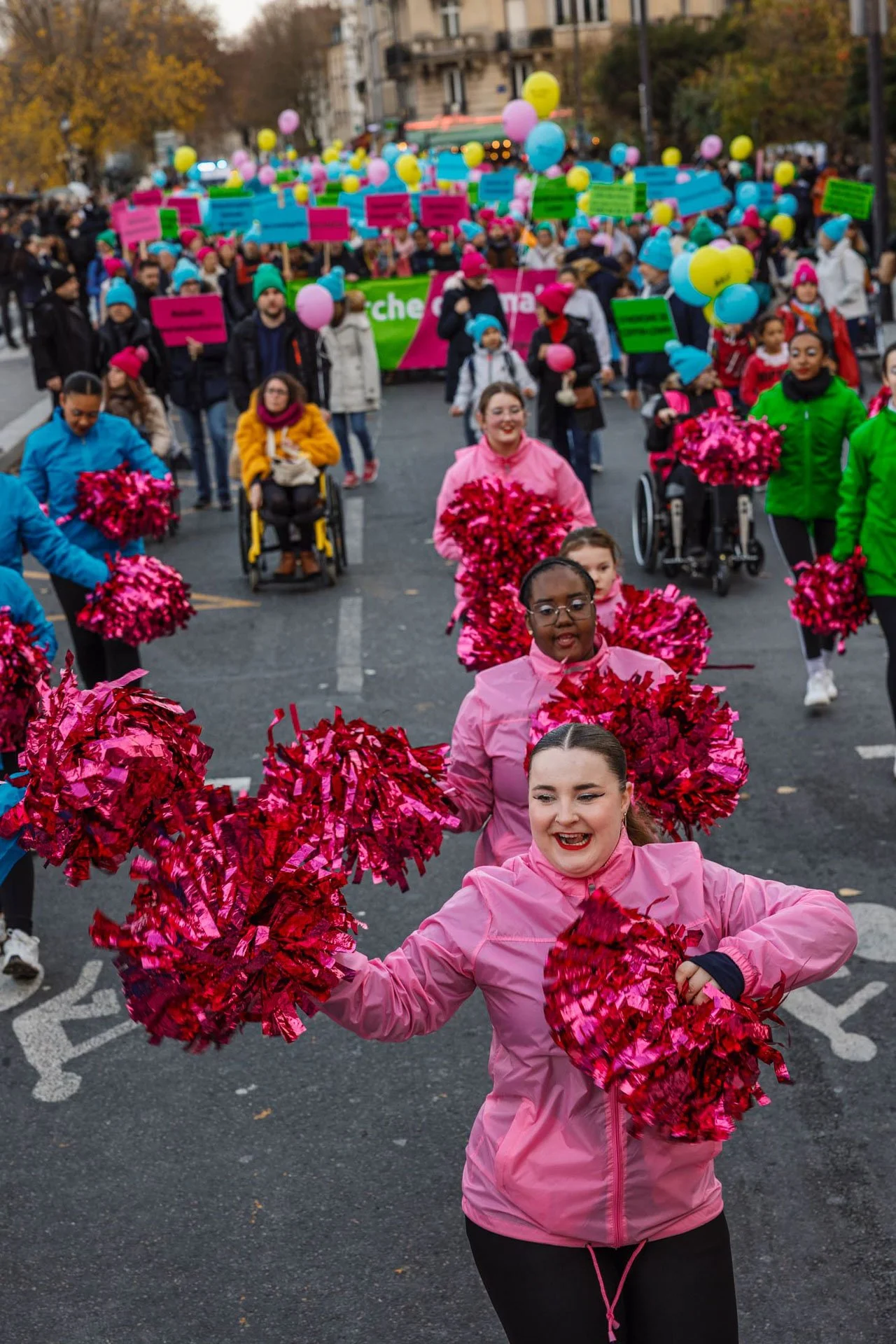 Un groupe de personnes, dont certaines en fauteuils roulants, participent à une parade ou un événement festif dans la rue, portant des vêtements colorés et tenant des pompons roses, avec des ballons colorés en arrière-plan.