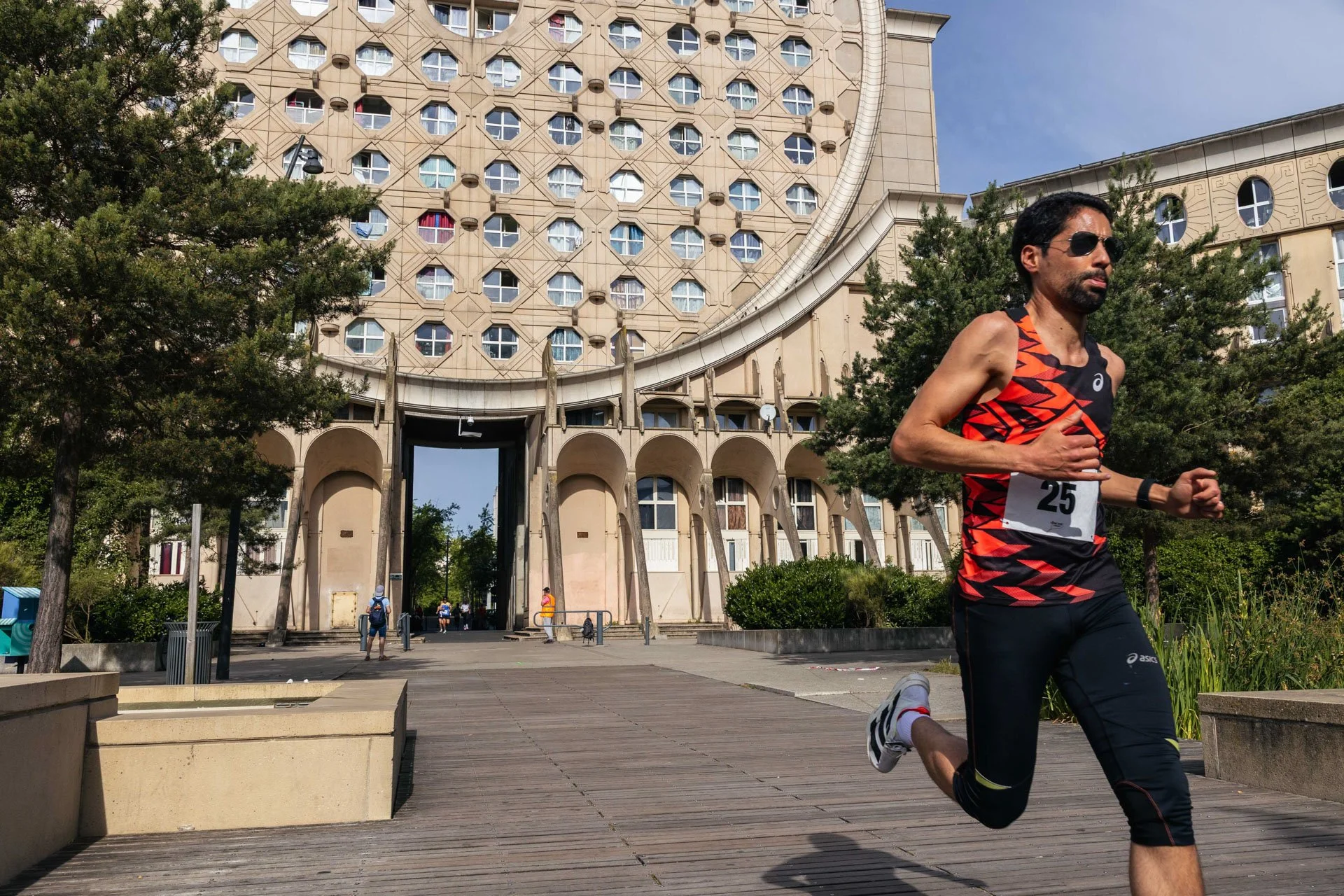 Un homme court dans une course, portant un maillot de sport noir et rouge numéroté 25, avec des bâtiments en arrière-plan et des arbres, sous un ciel en partie nuageux.