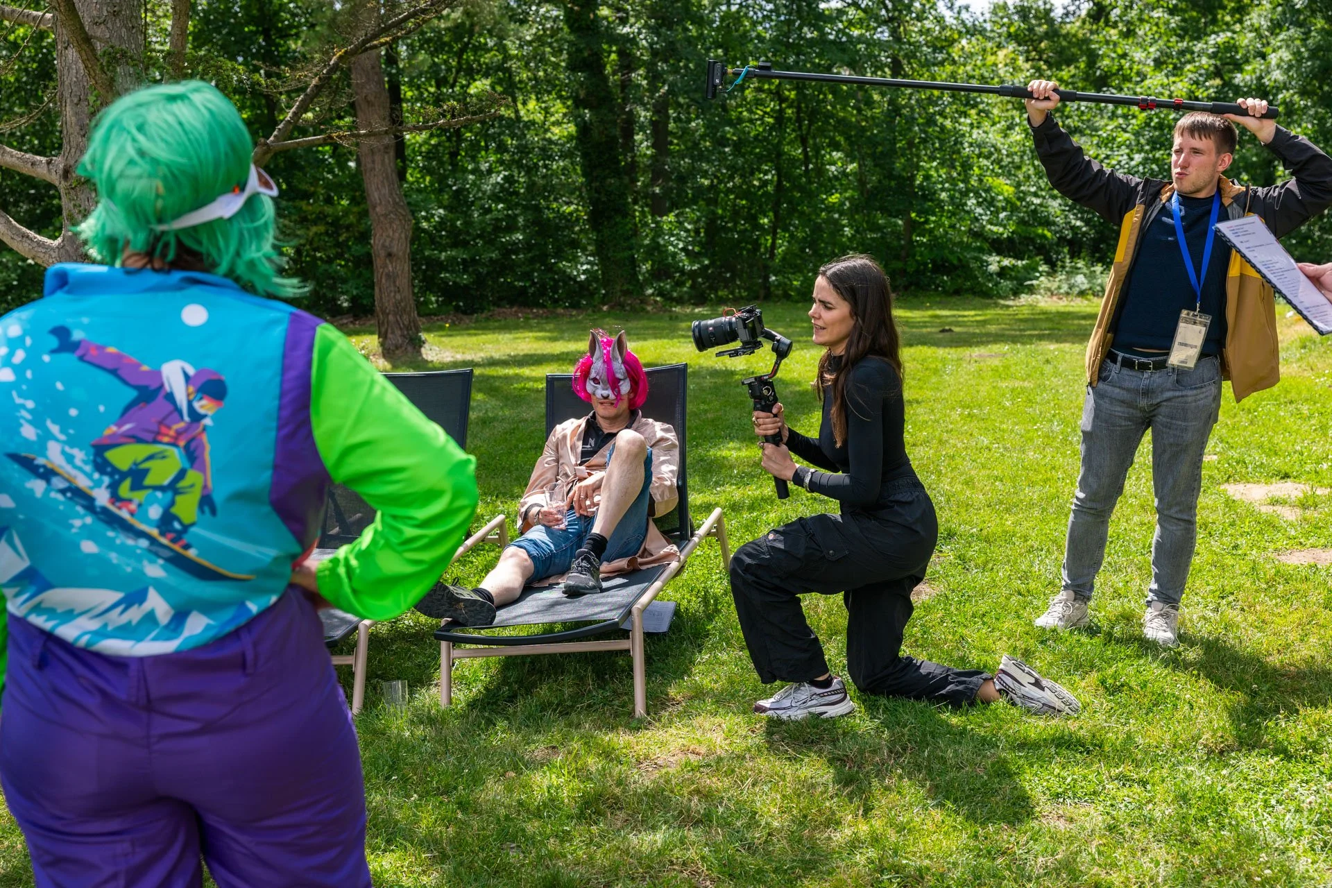 Groupe de personnes en train de filmer et photographier une personne portant un costume de licorne dans un parc ensoleillé. Une personne avec des cheveux verts est vue de dos, un homme avec bâton de micro et une femme avec un appareil photo stabilisa
