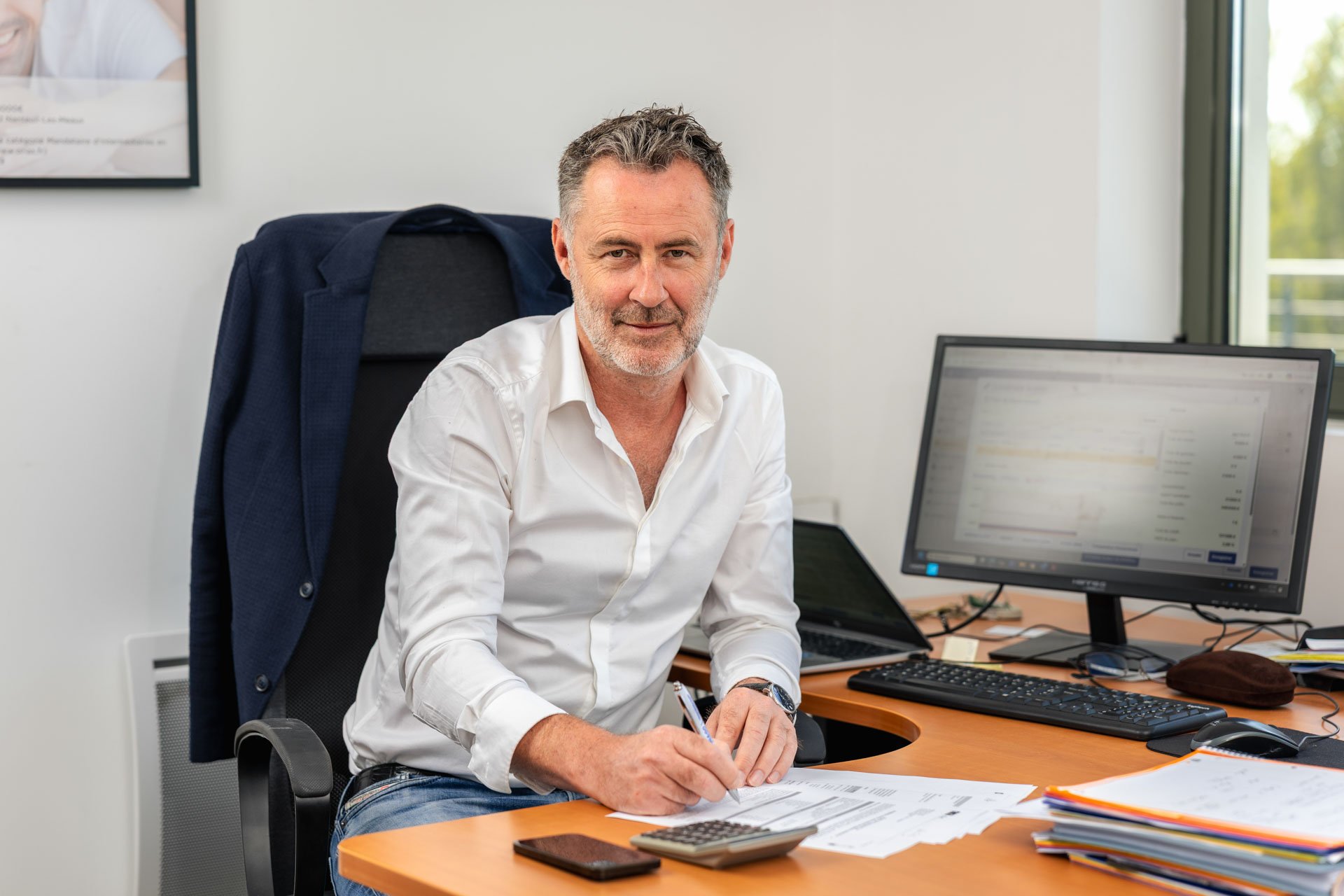 Homme d'affaires assis à un bureau, écrivant sur des papiers avec un ordinateur et une grande pile de documents en face, dans un bureau lumineux avec une fenêtre et un écran d'ordinateur.