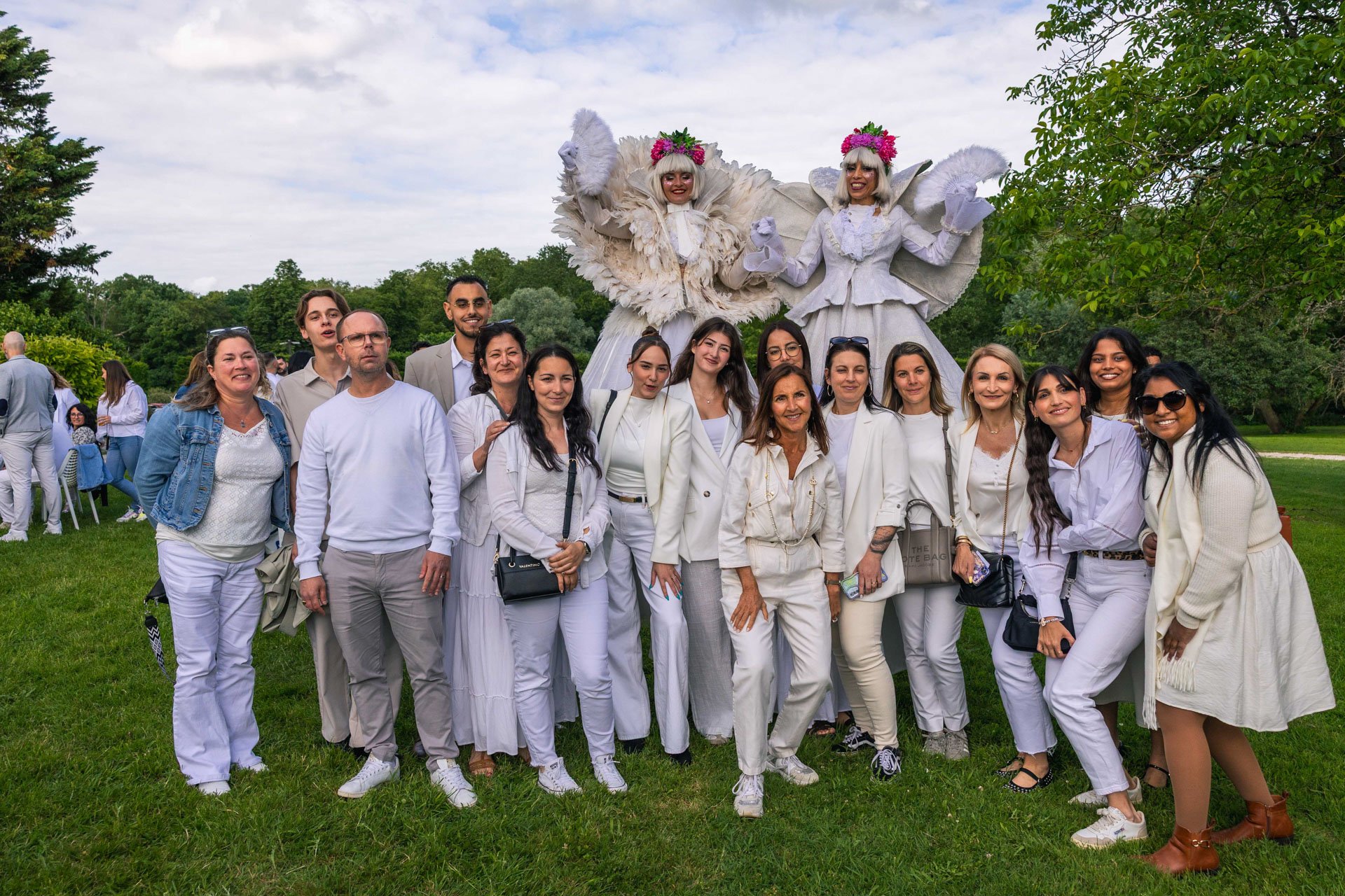Groupe de personnes posant pour une photo dans un parc, avec deux grandes figures de marionnettes ou costumes en arrière-plan représentant des anges blancs. La majorité des personnes portent des vêtements blancs ou neutres, et le ciel est partielleme