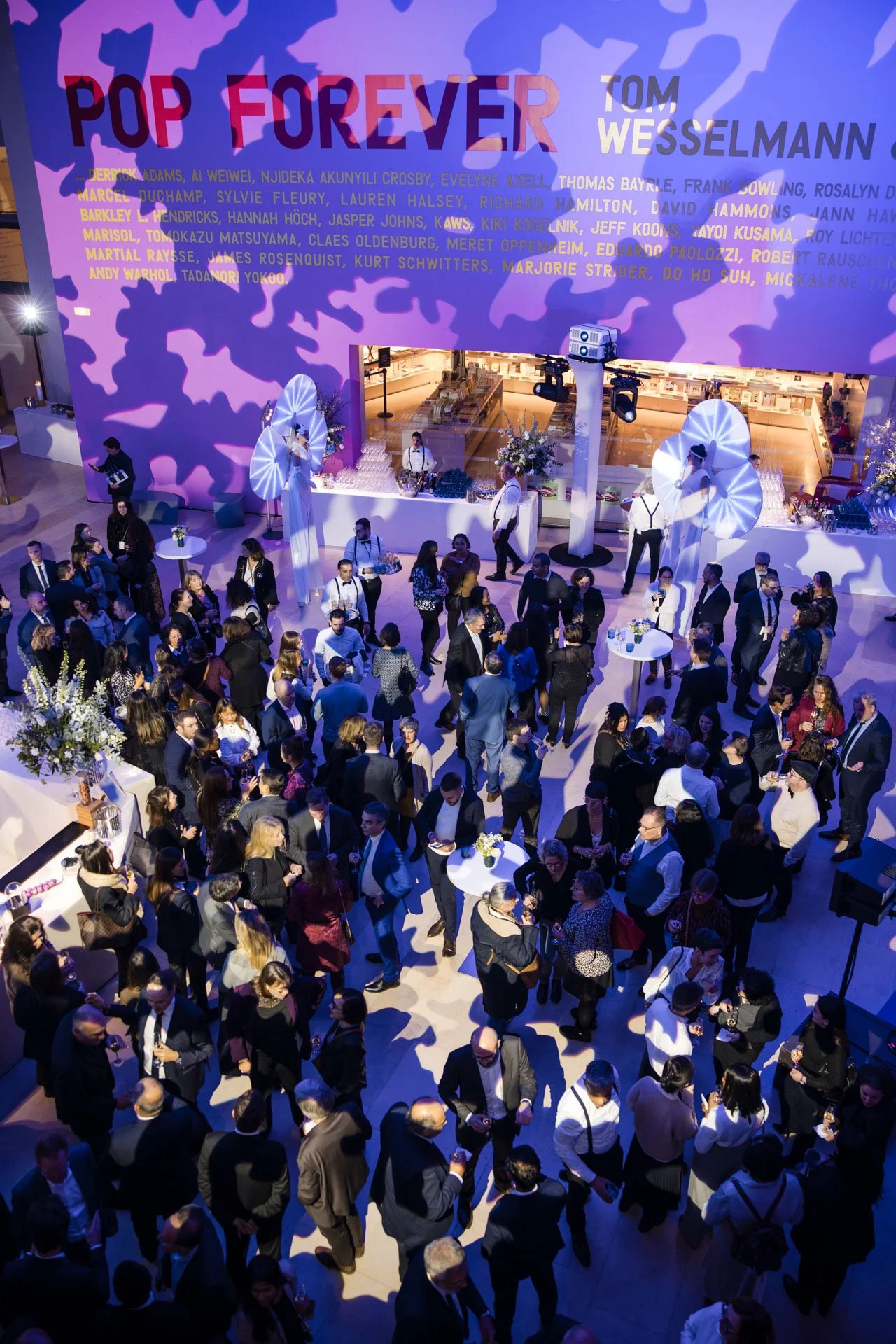 Une grande foule de personnes habillées en tenue formelle à un événement ou un cocktail dans une salle élégante, avec un grand écran au mur affichant de l'art graphique et des noms de célébrités ou d'invités.