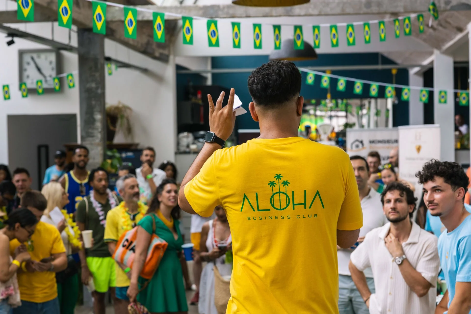 Un groupe de personnes participant à un événement social, décoré avec des drapeaux du Brésil. Au centre, une personne en t-shirt jaune avec le mot « ALOHA » et un palmier, vue de dos, semble organiser ou diriger le groupe.