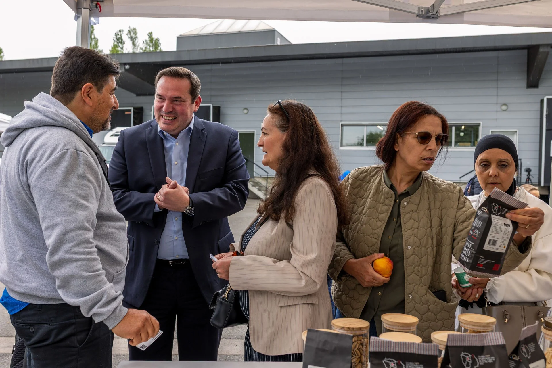 Groupe de cinq personnes discutant lors d'un marché ou stand en extérieur, avec en avant un comptoir rempli de bocaux et produits en emballages alimentaires.
