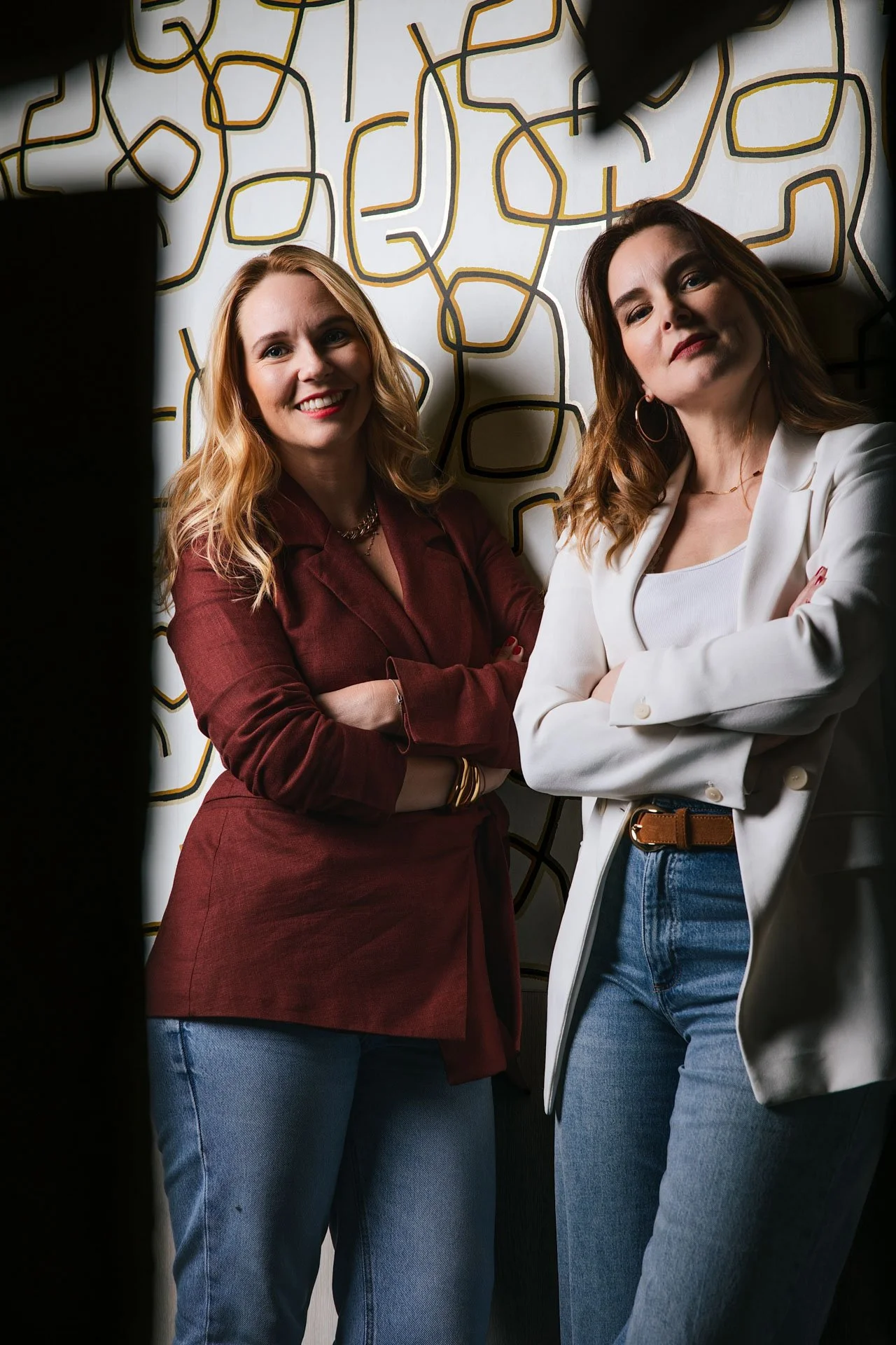 Deux femmes debout côte à côte avec leur bras croisés, souriantes, posant devant un mur à motif abstrait noir, blanc et jaune.
