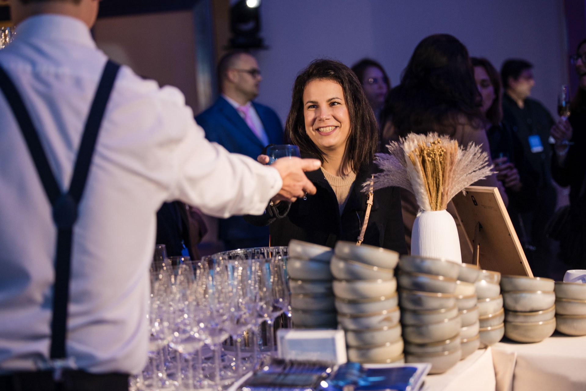 Une femme souriante en train de se faire servir un verre par un homme lors d'un événement social avec plusieurs personnes en arrière-plan.